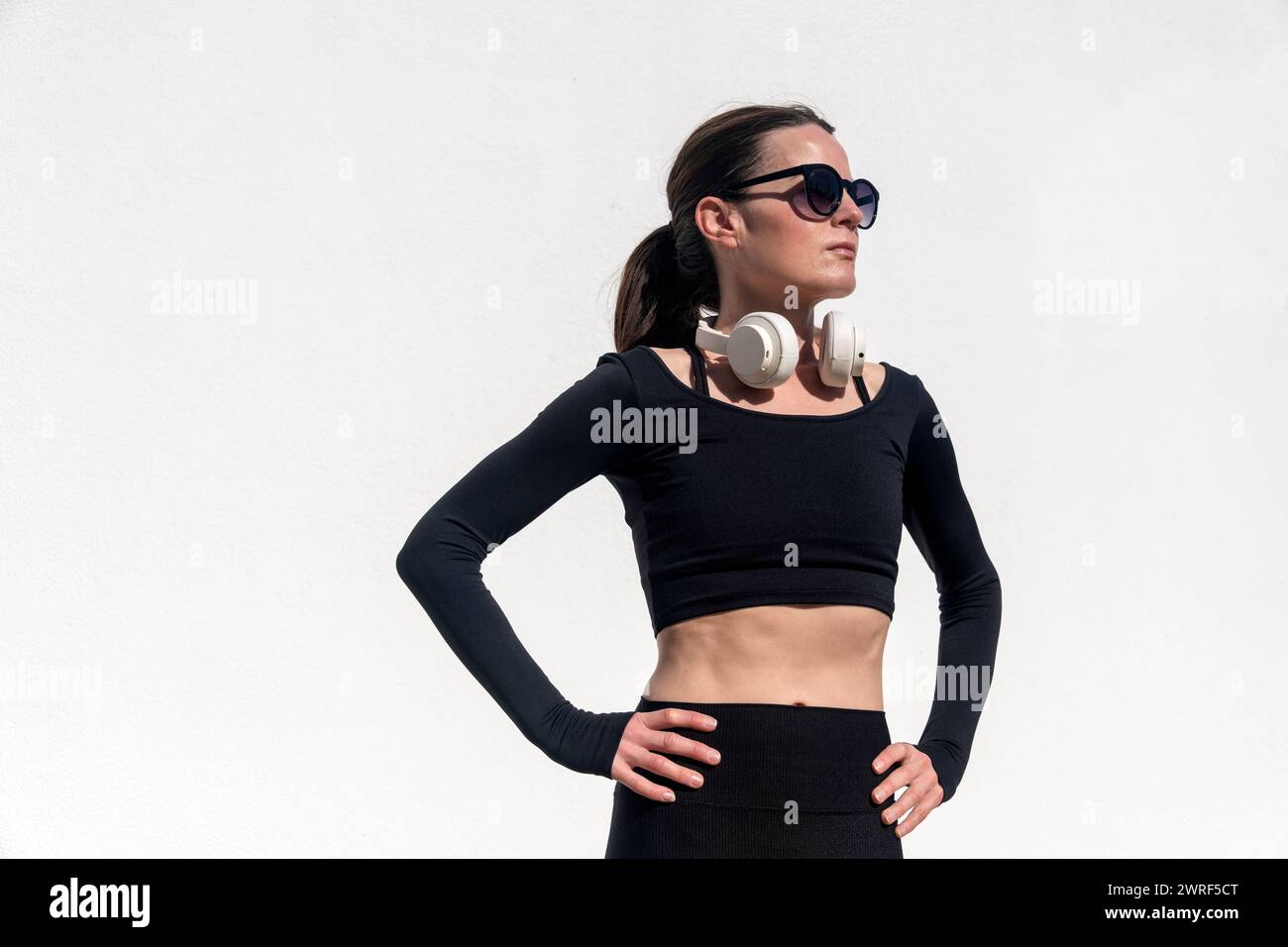 sporty woman with hands on her hips and headphones resting after exercise outdoors in the sun Stock Photo