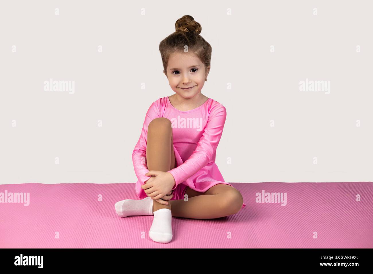 Portrait of a little gymnast girl in a pink dress and a bun is sitting on a pink fitness mat with her legs by her side and smiling beautifully at the Stock Photo