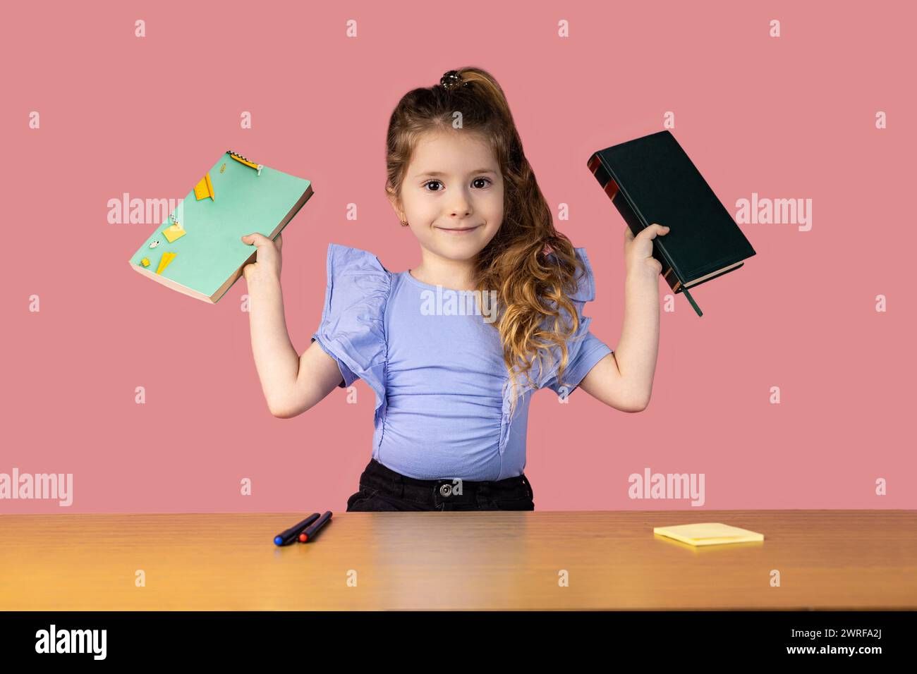 A little girl caught in a school desk holding two books showing interesting emotions, the girl loves to read books and go to school. High quality phot Stock Photo