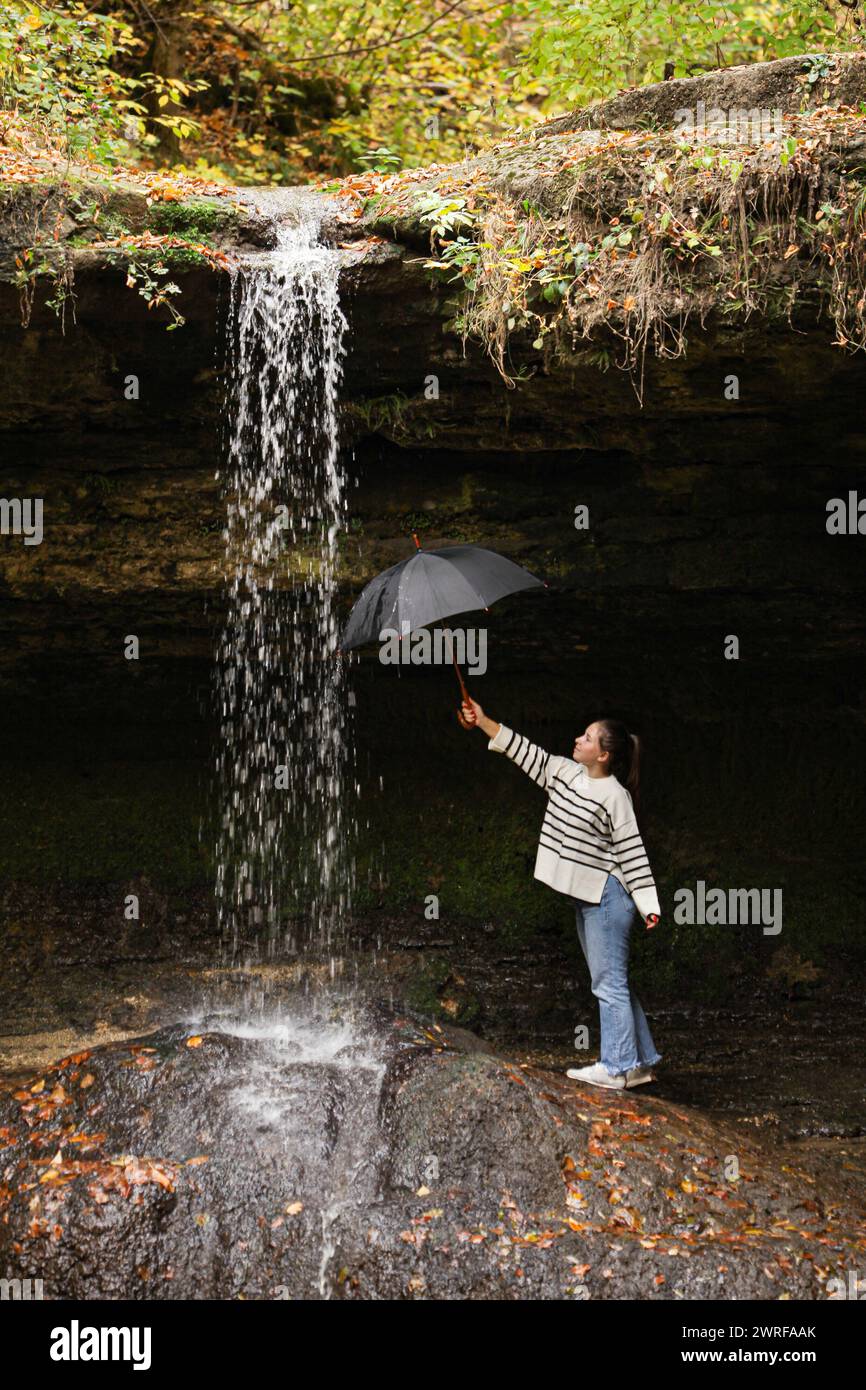 young girl standing by the waterfall flowing streams in the morning from a cave low angle holding a black umbrella under the flowing water of the big Stock Photo