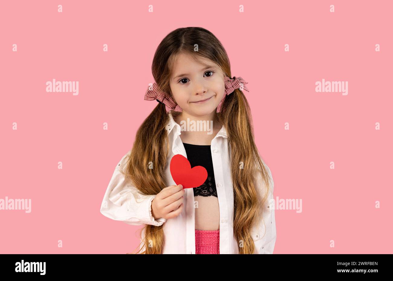 A little girl with two pigtails caught with two pink tops, who is pictured on a pink background holding a heart, is dedicated to her mother or to all Stock Photo