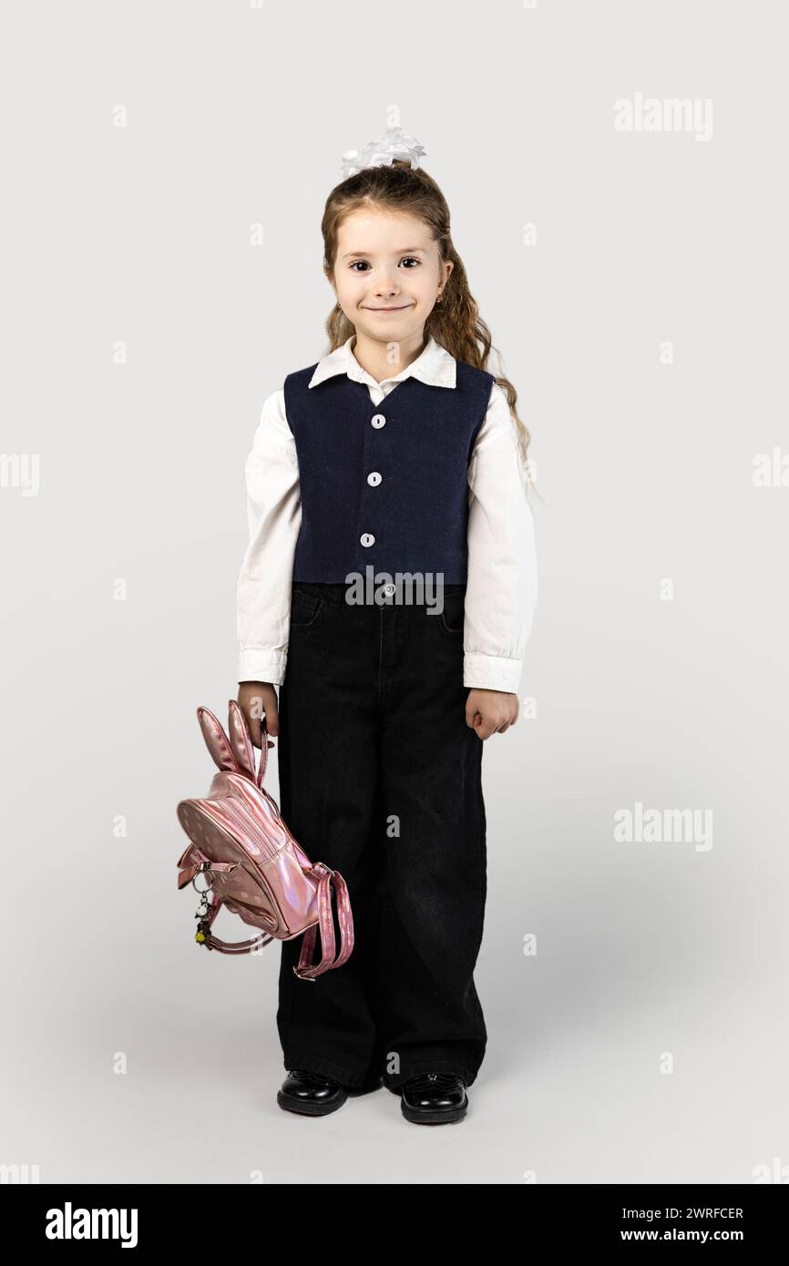 A little girl in a school uniform with an electric blue blazer and collar is holding a pink backpack. She smiles brightly, her thumb hooked through th Stock Photo