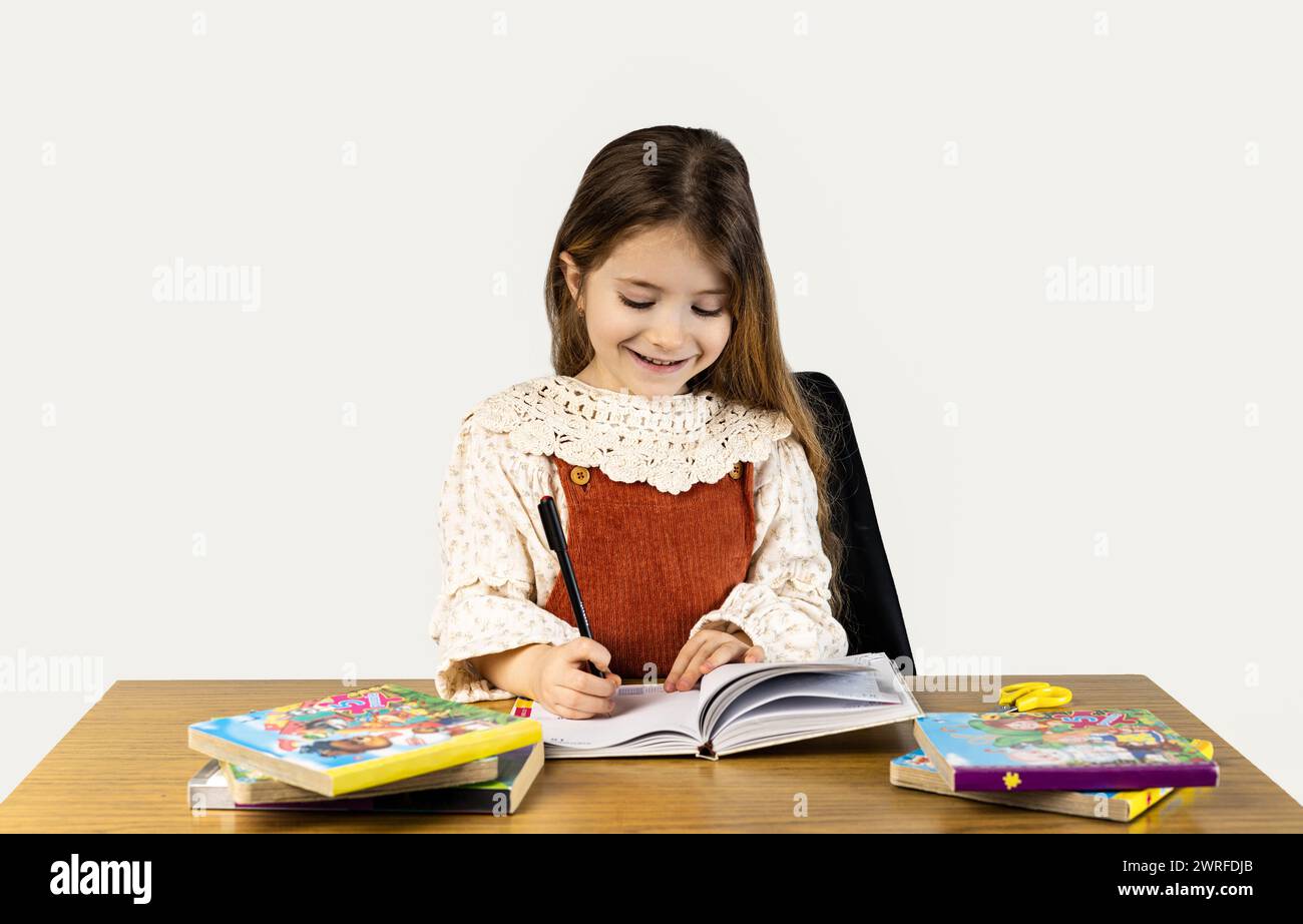 A happy little girl is sitting at a table, smiling while reading a book. Her sleeves are rolled up as she enjoys leisure time, sharing the desk with o Stock Photo