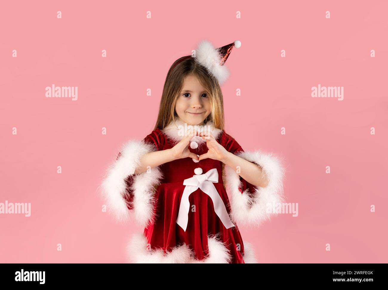 An attractive little girl with blond hair, dressed in a Christmas dress, on a pink background shows her heart with her hands. High quality photo Stock Photo