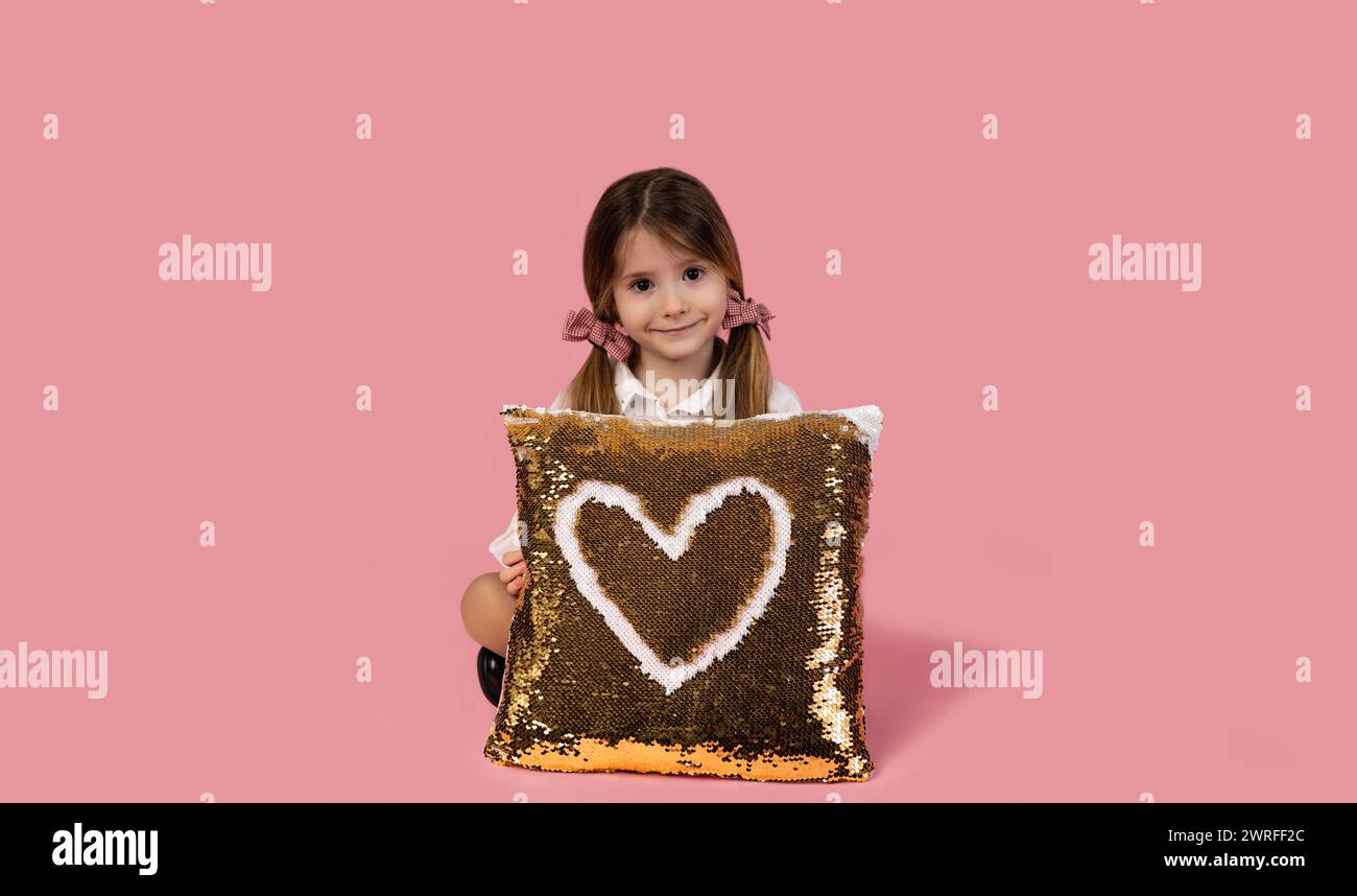 Stylish little girl with two ponytails standing isolated on pink background holding a golden sequin pillow with a heart drawn on it, Valentine's day c Stock Photo