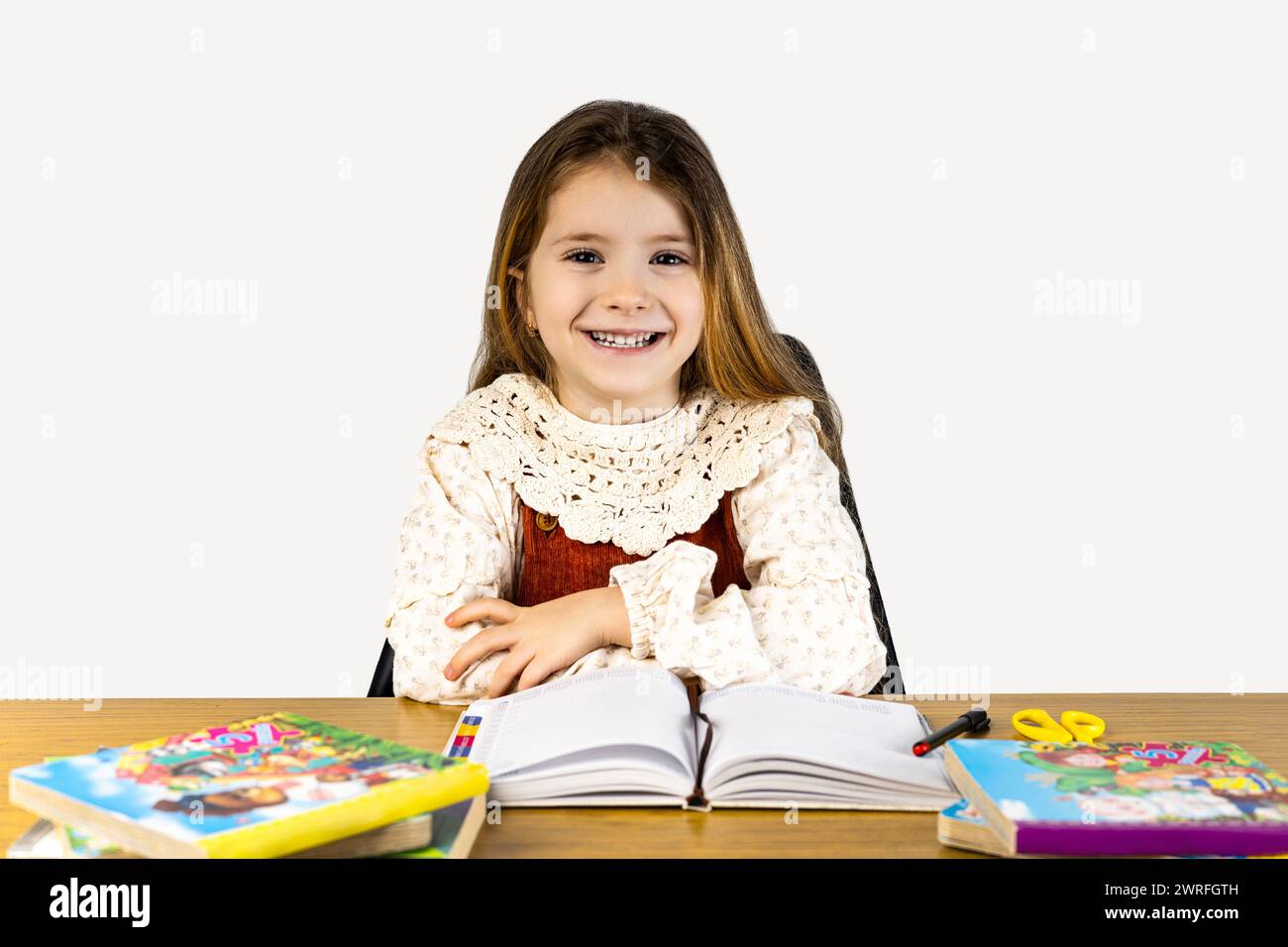 A happy toddler is sitting at a desk, sharing books and having fun. Her smile lights up the room as she leisurely flips through the pages Stock Photo