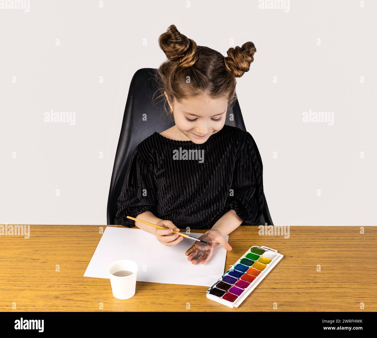 A little girl is sitting at a hardwood table painting with watercolors ...