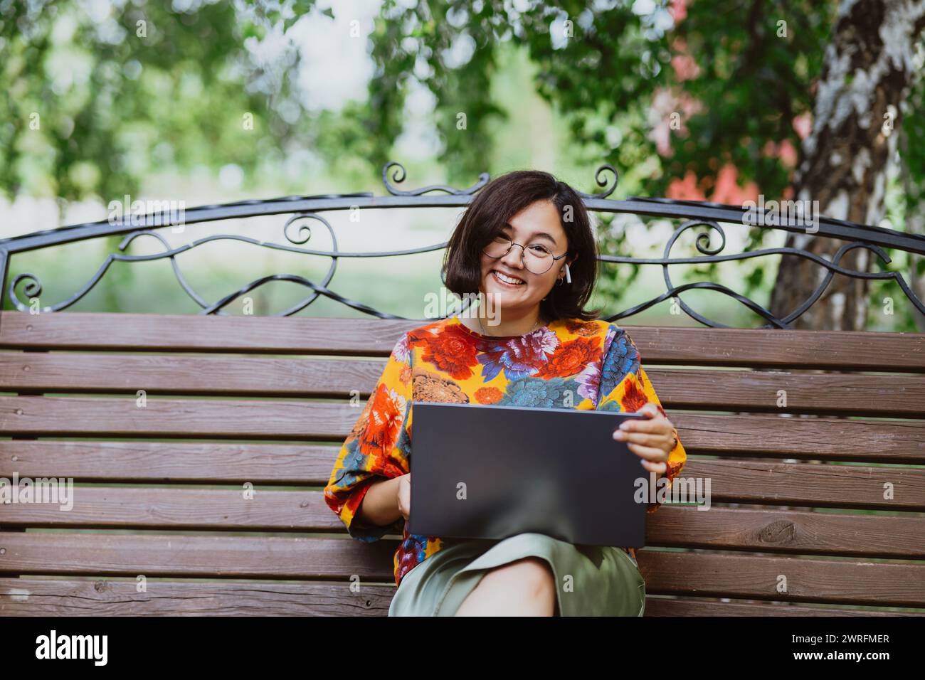 Smiling Asian woman working on her laptop while sitting on a park bench ...
