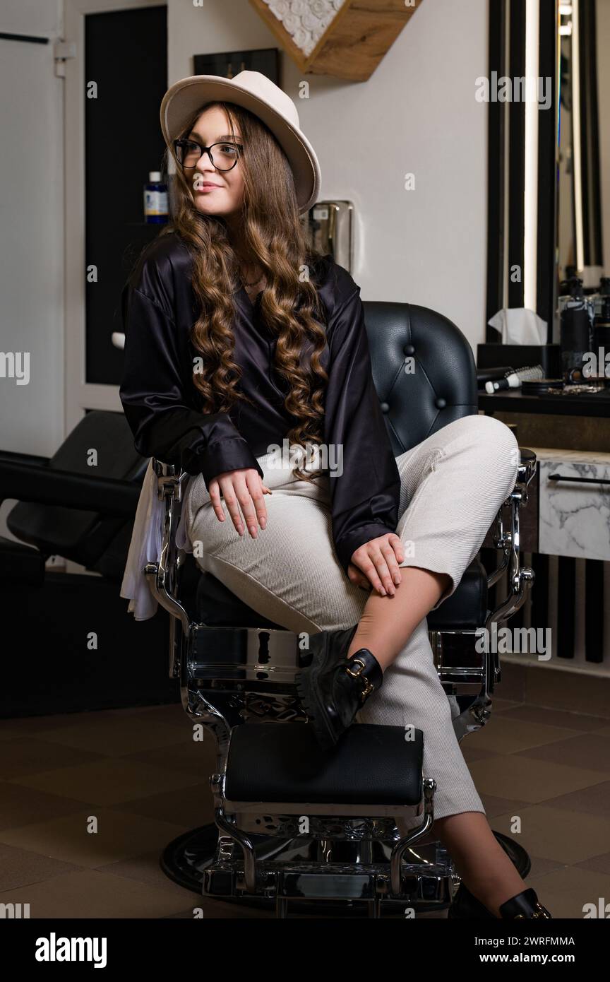 A female barber is sitting in a chair and waiting for a client. Portrait of a young hairdresser girl in a hat. Stock Photo