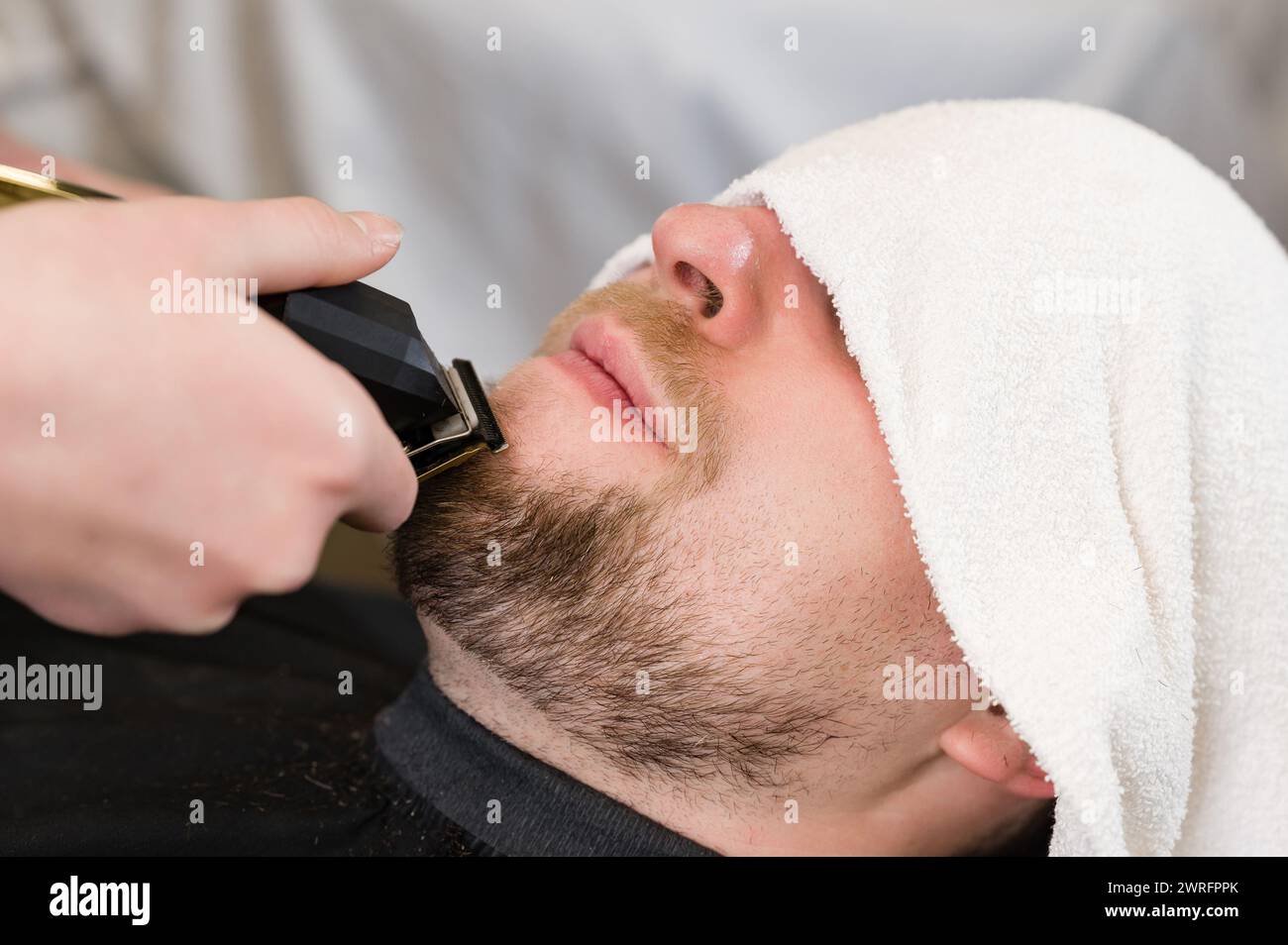 The client's head is covered with a towel during a beard trim. Shortening the length of the beard from the sides by the master for the client. Stock Photo