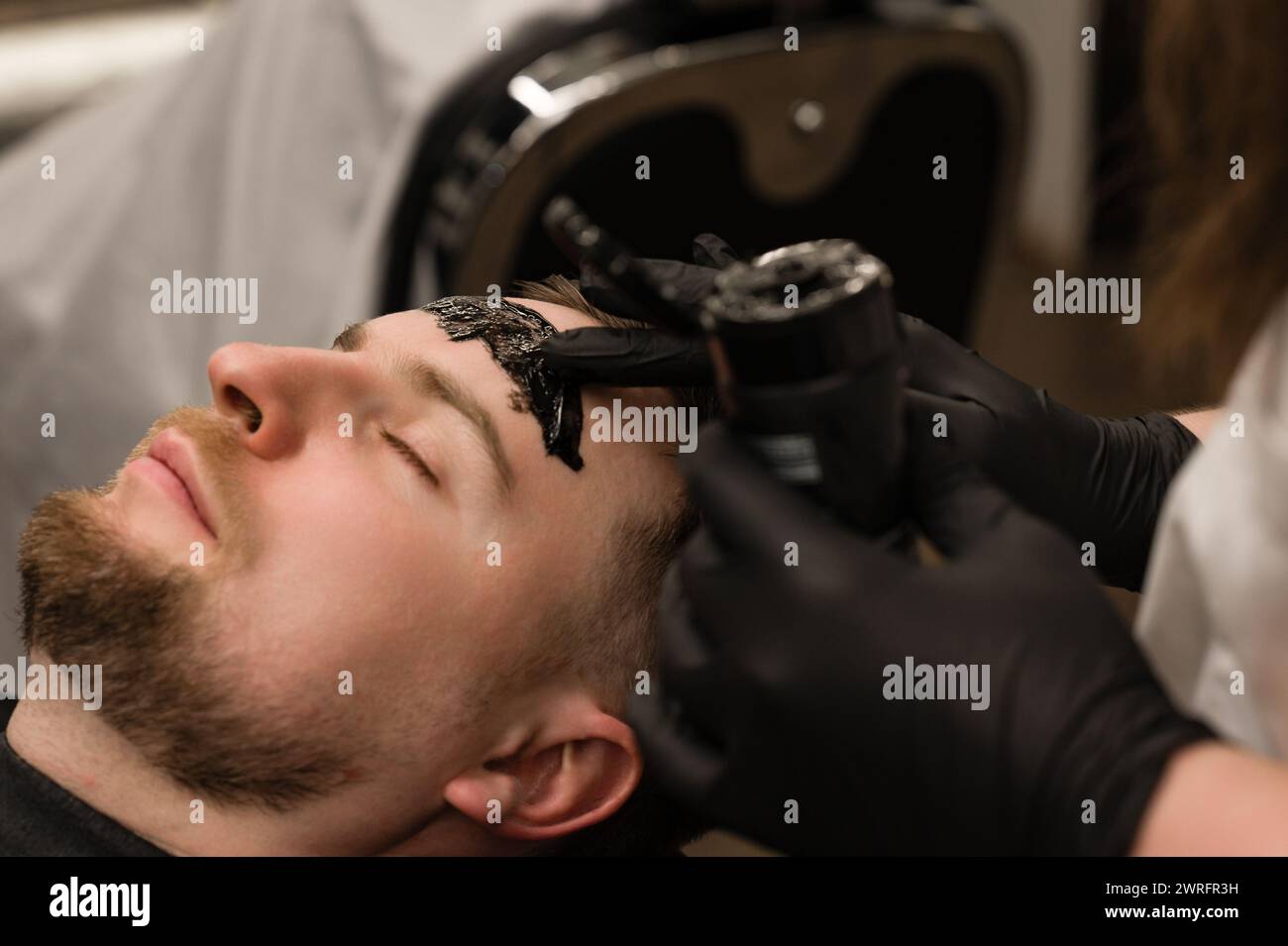 Barber applies a black moisturizing mask to a mans face while shaving. A dermatologist applies a black mask to the face of a bearded patient to clear Stock Photo