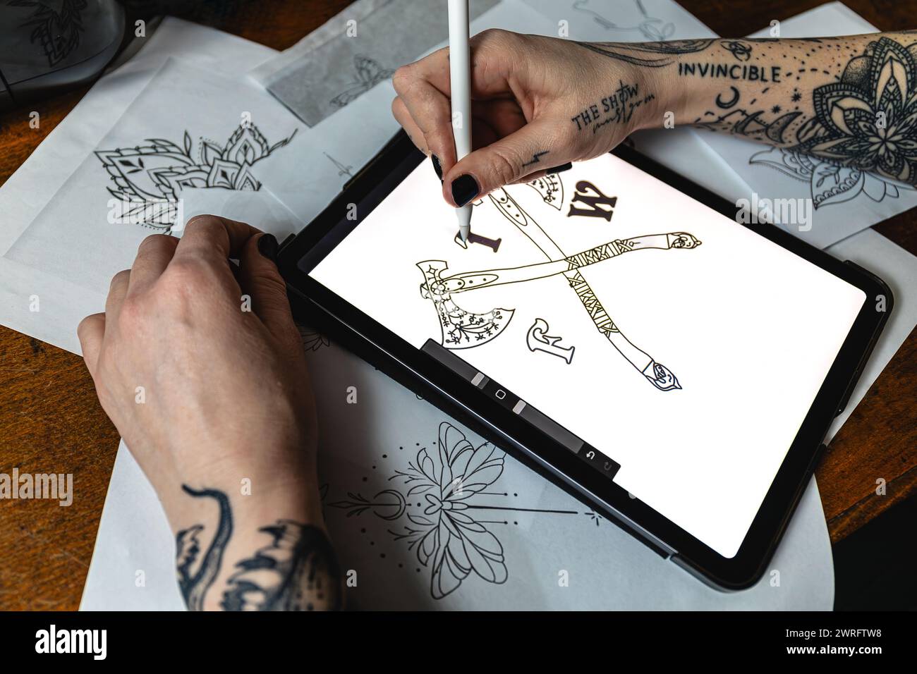 Horizontal photo an artist's hand carefully renders a tattoo design on a graphic tablet amid a spread of paper sketches. Concept business, art. Stock Photo