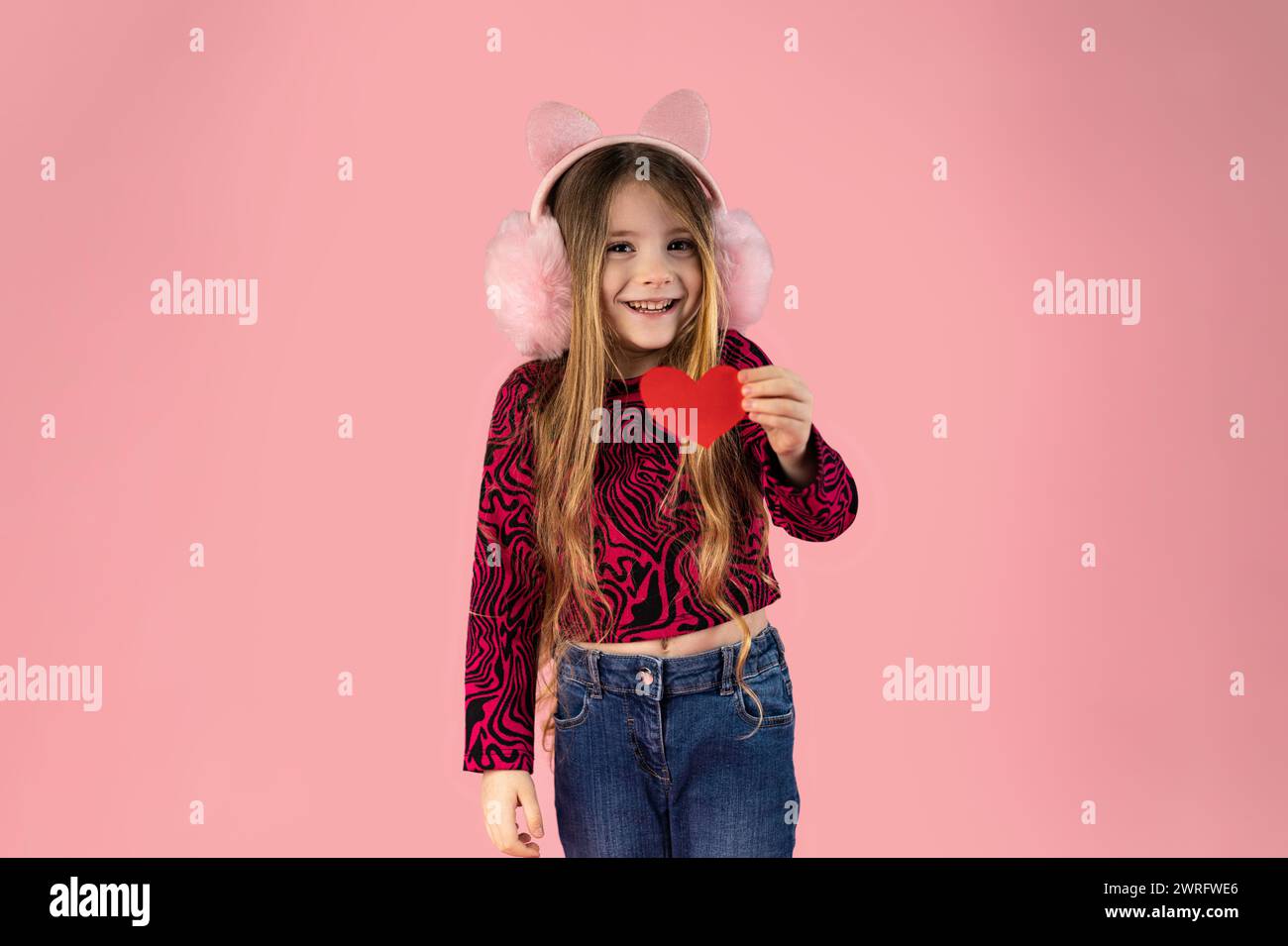 Little girl with long blond hair wearing pink headphones on her ears, dressed in jeans and a pink printed sweater holds a small paper heart and smiles Stock Photo