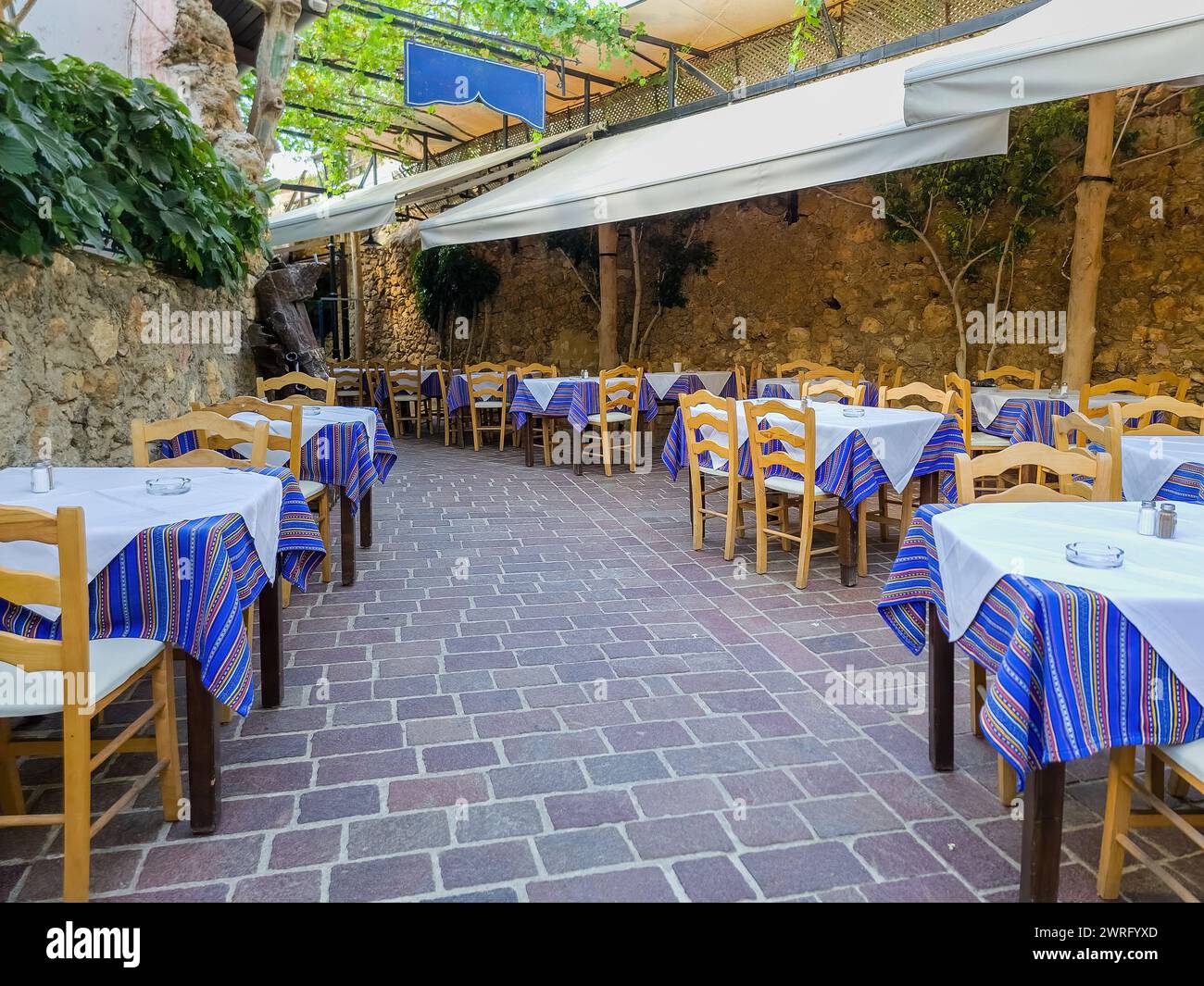 Open air traditional taverna at Chania Old Town, afternoon in Crete island Greece. Tavern restaurant, empty chair table, paved alley. Stock Photo