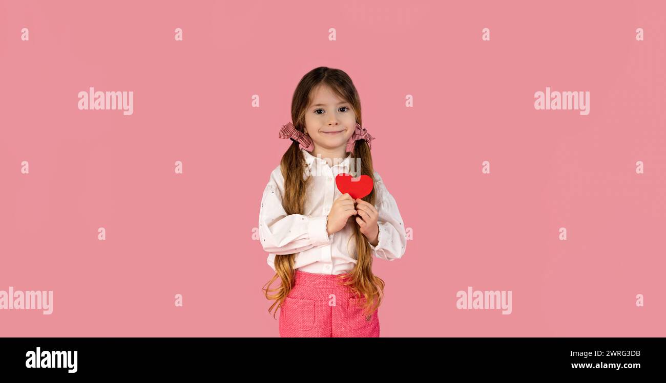 The emotional blonde, stylish little girl with two pigtails is holding a red paper heart on her chest on a pastel pink background, Valentine's day con Stock Photo