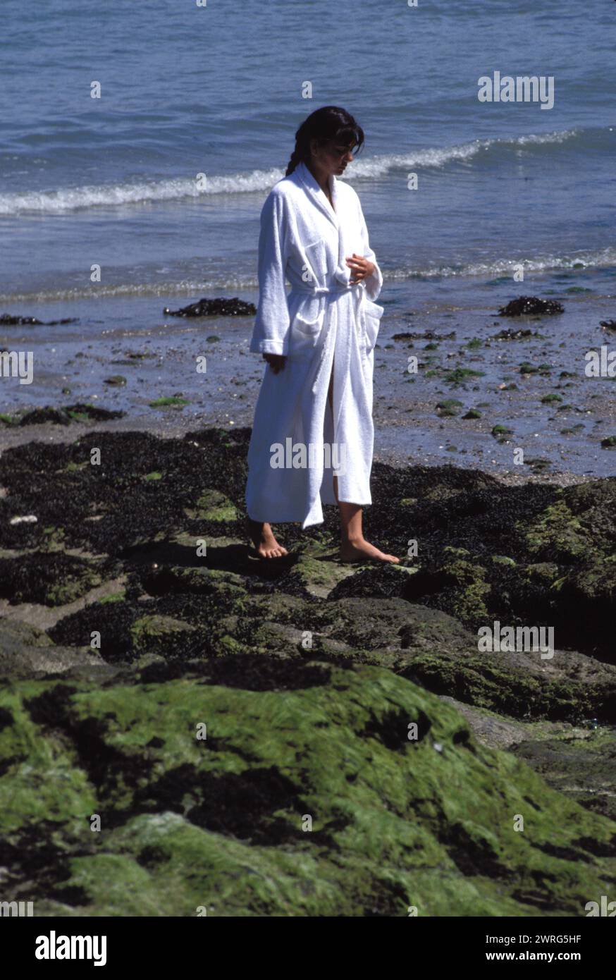 black hair young woman st malo france spa resort thalassotherapy walking on the shore with seagrass in white bath dress Stock Photo