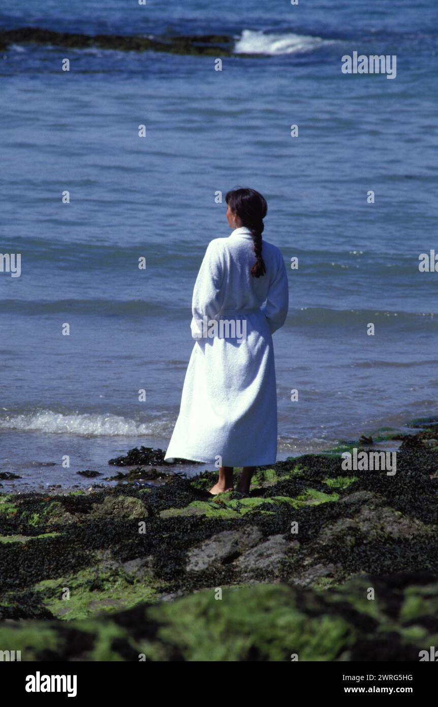 black hair young woman st malo france spa resort thalassotherapy walking on the shore with seagrass in white bath dress Stock Photo