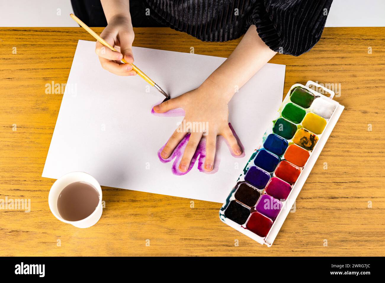A person is painting a hand with watercolors on a piece of paper placed on a hardwood table. The paintbrush is held delicately between their fingers Stock Photo