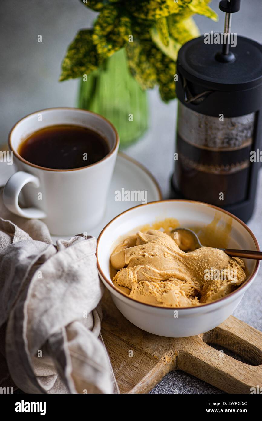 Close-up of a cup of black coffee with a bowl of caramel ice cream on a wooden chopping board with a cafetiere and foliage Stock Photo
