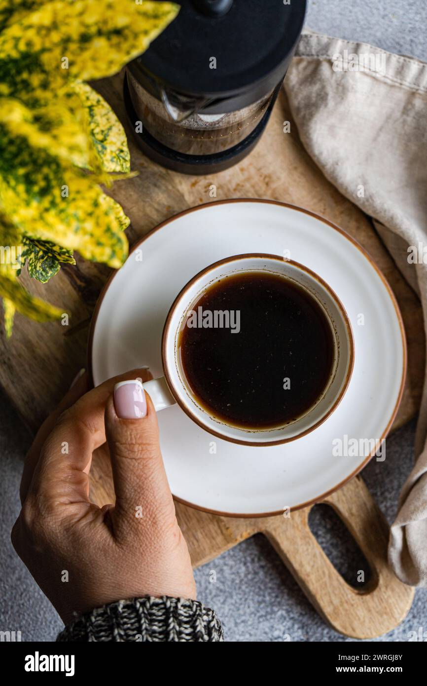 Overhead view of a Woman reaching for a cup of black coffee on a chopping board with a cafetiere, napkin and foliage Stock Photo