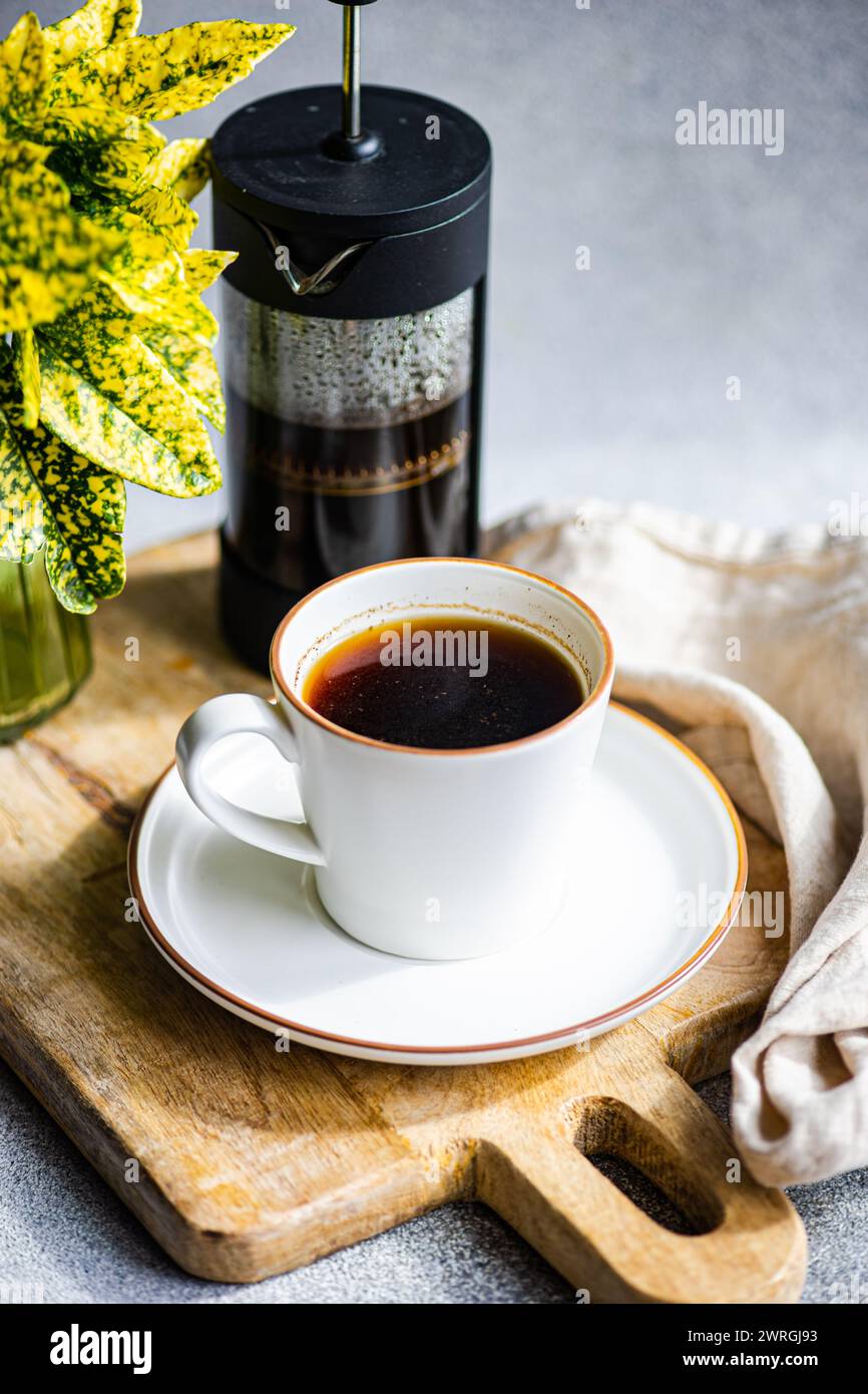 Close-up of a cup of black coffee on a chopping board with a cafetiere, napkin and foliage Stock Photo