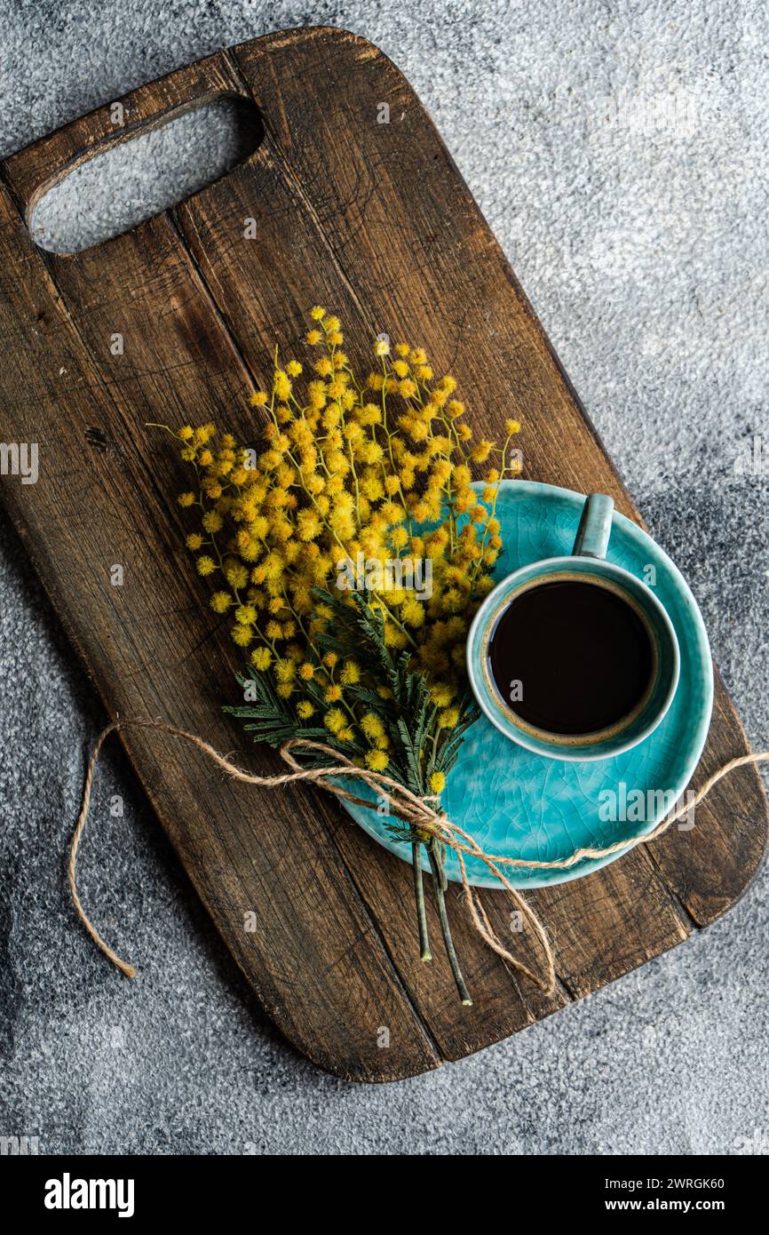 Overhead view of a cup of black coffee and a bunch of yellow mimosa flowers on a wooden chopping board Stock Photo