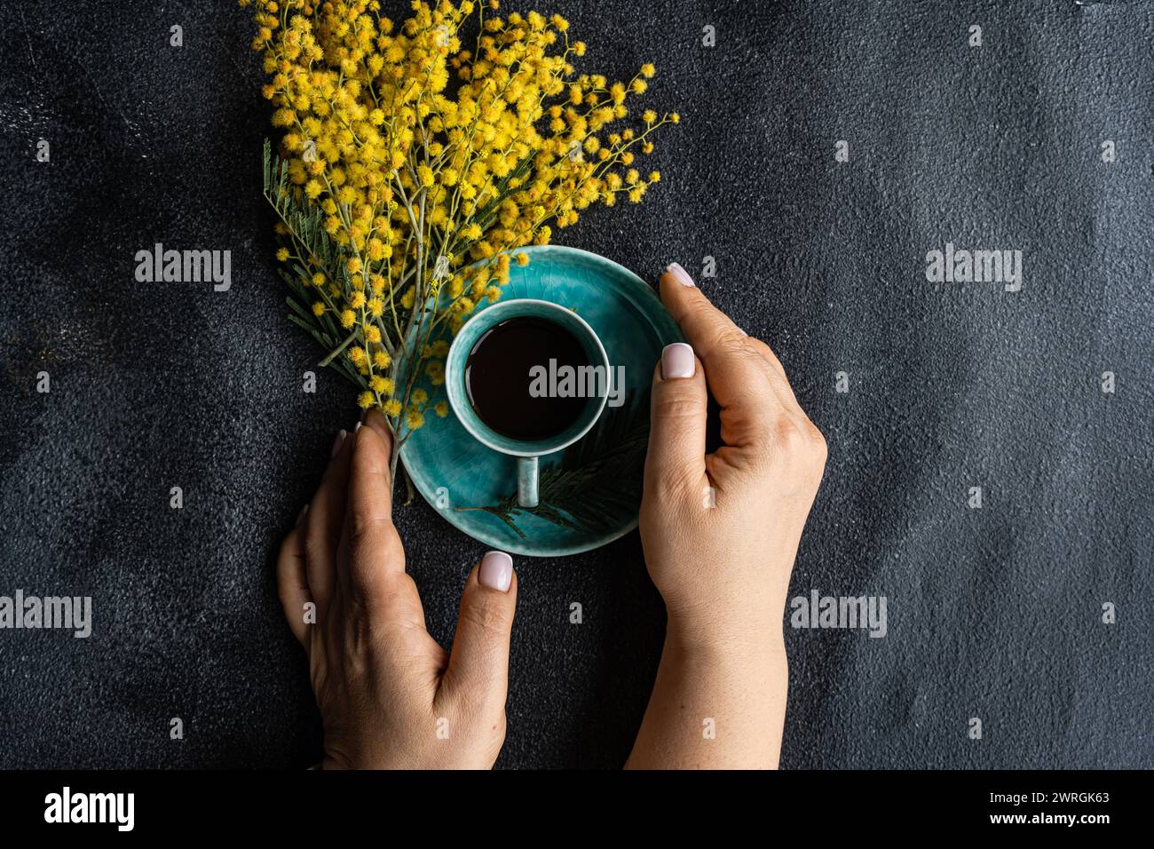 Overhead view of a woman reaching for a cup of black coffee next to a bunch of yellow mimosa flowers on a table Stock Photo
