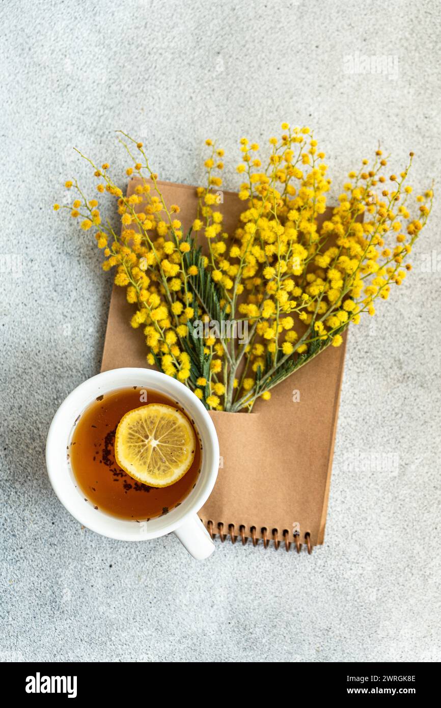 Overhead view of a cup of tea with lemon and a bunch of yellow mimosa flowers tucked into a notepad on a table Stock Photo