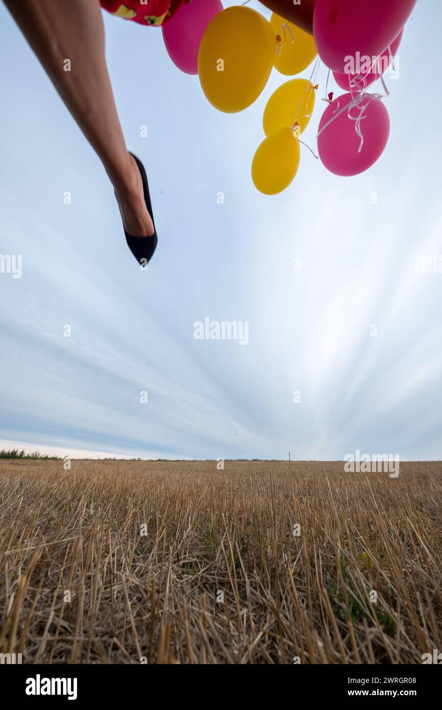 shot from ground below to foot part of a woman with yellow and red balloons running and jumping outdoors in the fields, copy space Stock Photo