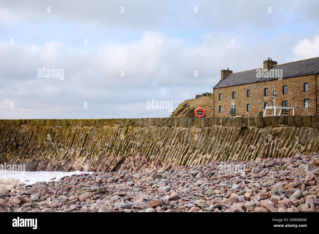 Stone cottage at Sinclairs Bay harbour entrance, with the sea rolling in on the stone shingle and fishing boats behind harbour wall Stock Photo