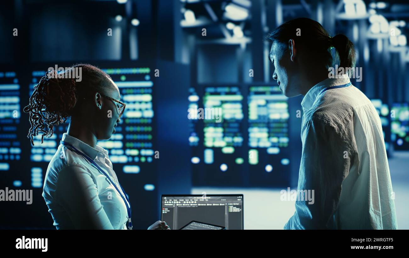 Technicians programming in server room housing advanced infrastructure storing massive datasets. Workers running code on laptop to upgrade data center racks doing complex computational operations Stock Photo