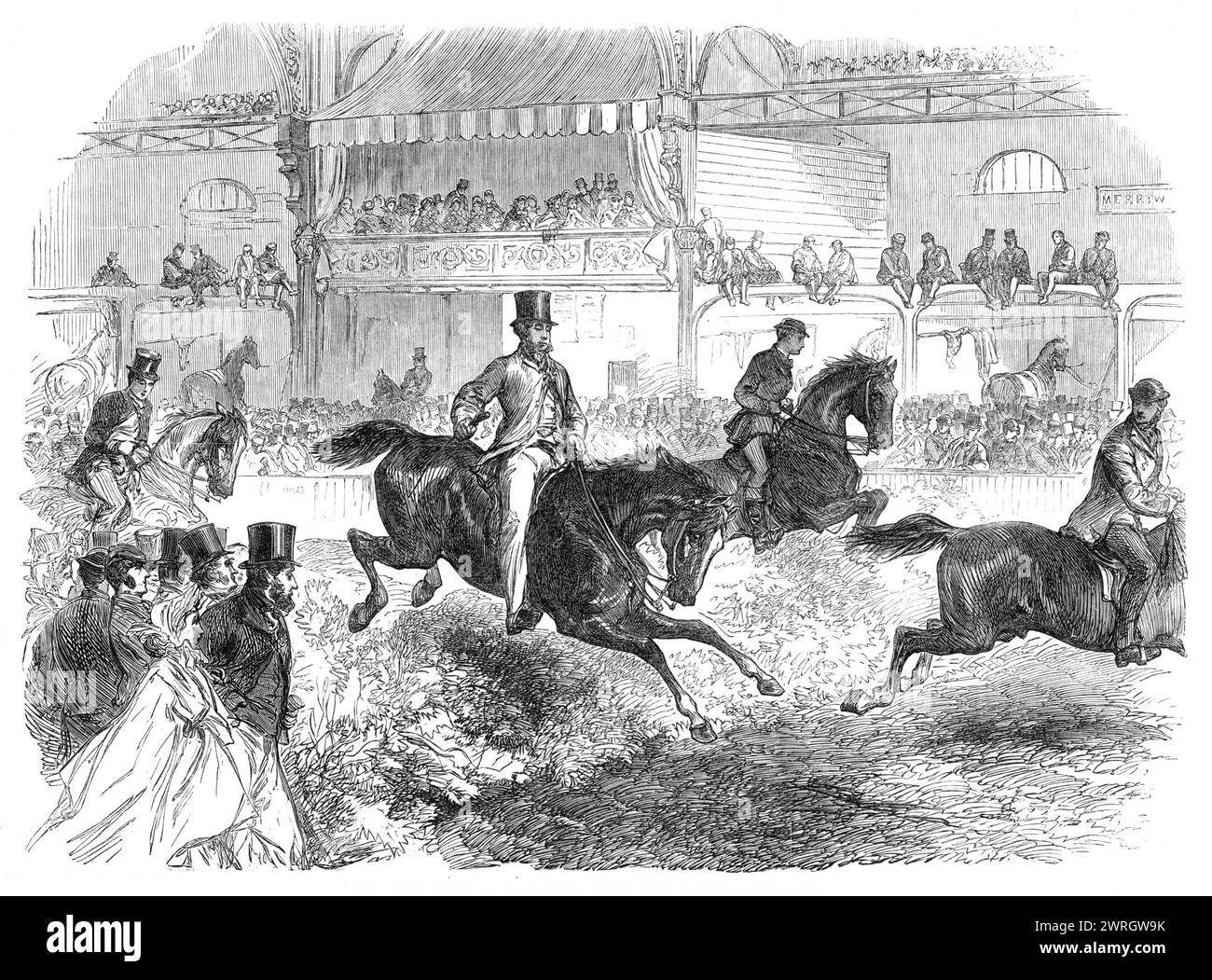 The Horse Show at Islington [in London]: trying the hunters, 1864. View of '...the Trial of Hunters jumping over a framework of hurdles covered with furze-bushes to represent a hedge. The hurdles were fixed upon an axis which would revolve at the least touch; so that there was no fear of throwing down the horse whose feet should strike against them. The central area of the hall had been inclosed and covered with tan, so as to form a safe and convenient circus for the performance of these equine feats, and for the promenade of the different classes of horses, either led, ridden, or driven - wal Stock Photo