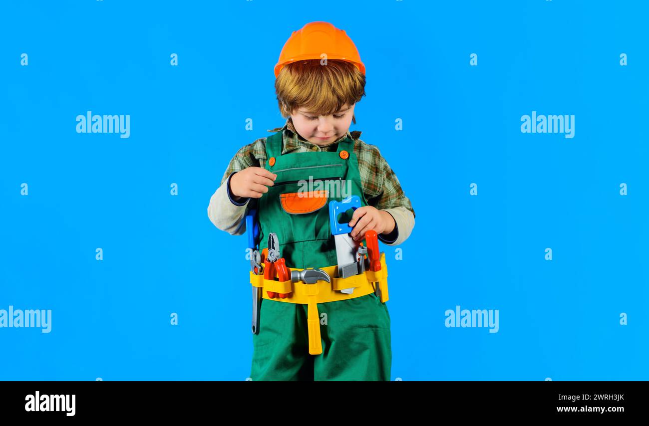 Little builder boy in helmet and toolbelt playing with toy saw. Child game. Cute kid in protective hard hat with tool belt of toy tools for repair Stock Photo