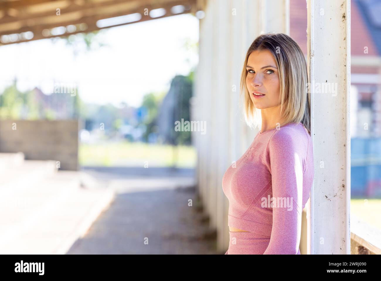 In the bright ambience of an urban space, a woman stands poised in form-fitting pink athletic attire, her focused expression capturing a moment of vigilance before activity. Urban Fitness Vigilance: Woman in Pink Athletic Attire Poised for Action. High quality photo Stock Photo