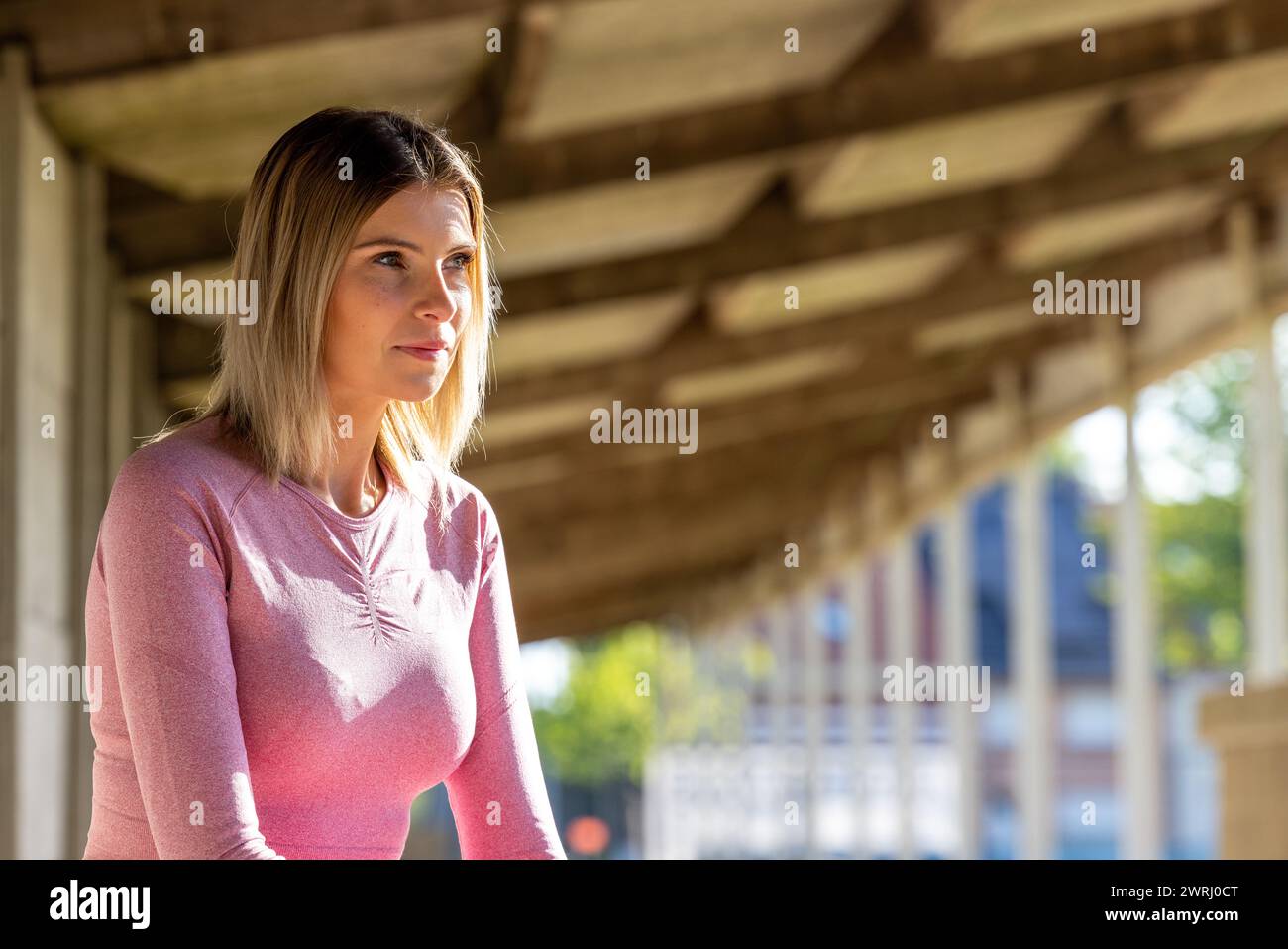 A young woman in a pink long-sleeve top and leggings stands in a shaded urban corridor, her attention captured by a distant point, reflecting a moment of calm before action. Reflective Moment: Woman in Pink Fitness Attire Gazing into Distance. High quality photo Stock Photo