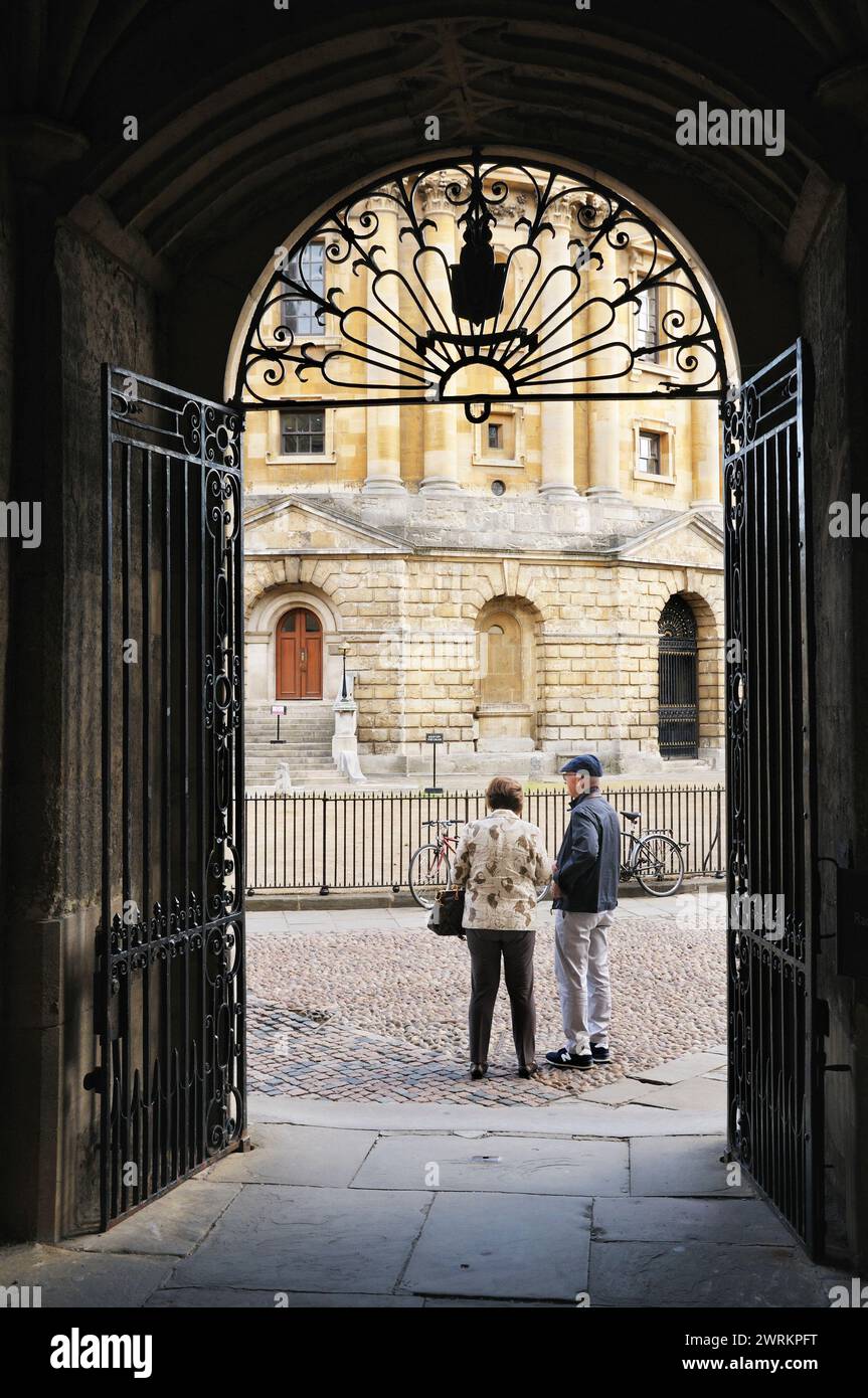 View through Bodleian Library wrought iron gate arch with senior couple standing in Radcliffe Square next to Radcliffe Camera, University of Oxford UK Stock Photo