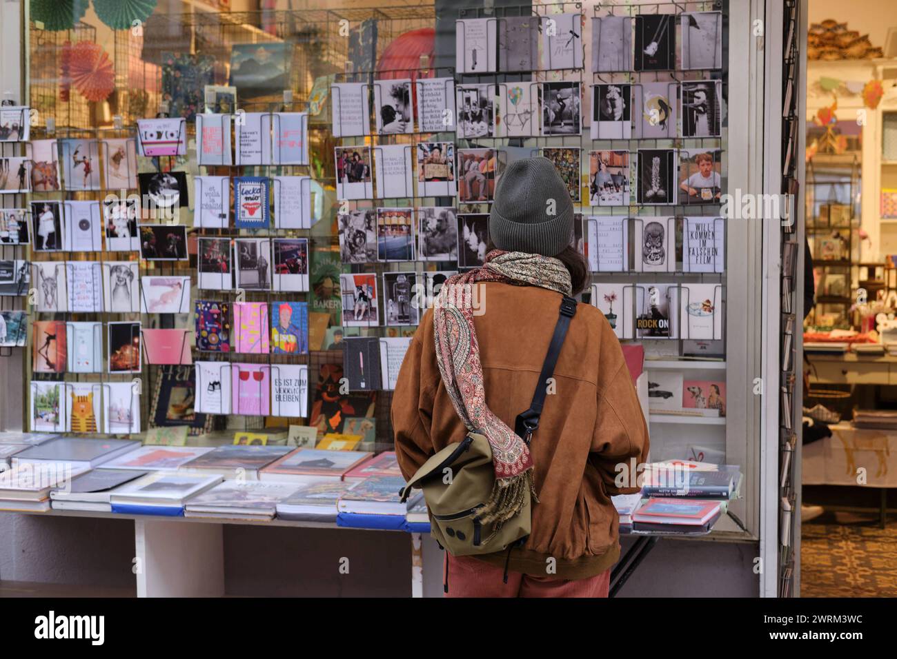 Junge Frau betrachtet Postkarten Junge Frau mit Mütze, Schal und Umhängetasche betrachtet Postkarten in Schaufenster. München *** Young woman looking Stock Photo
