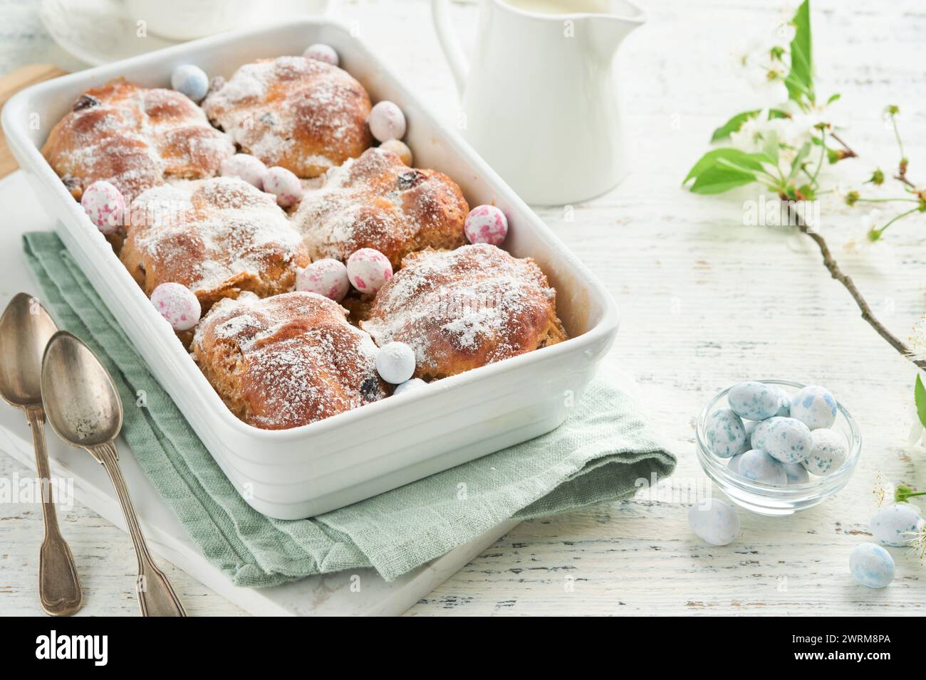 Easter Hot cross buns. Traditional Easter treats cross buns with raisins, butter, chocolate candy eggs with blossoming cherry or apple flowers on rust Stock Photo