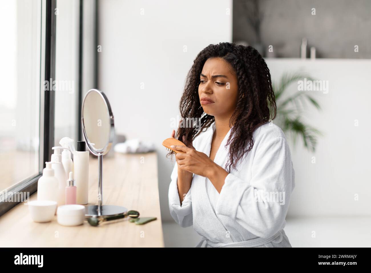 Worried black woman in bathrobe struggling with tangled hair in hairbrush Stock Photo