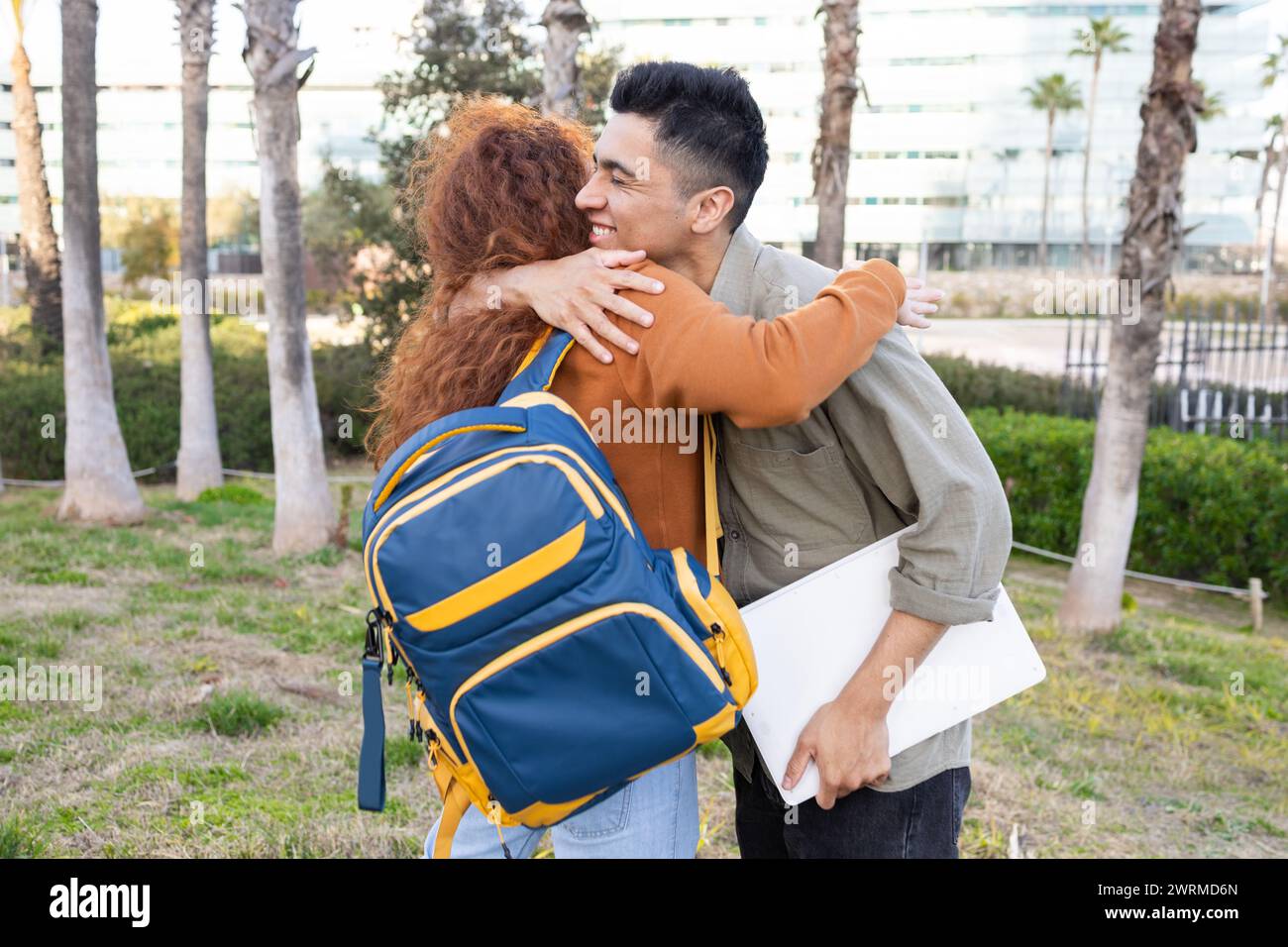 An affectionate hug between a woman and a man on a college campus, with the woman carrying a blue and yellow backpack and the man holding a white lapt Stock Photo