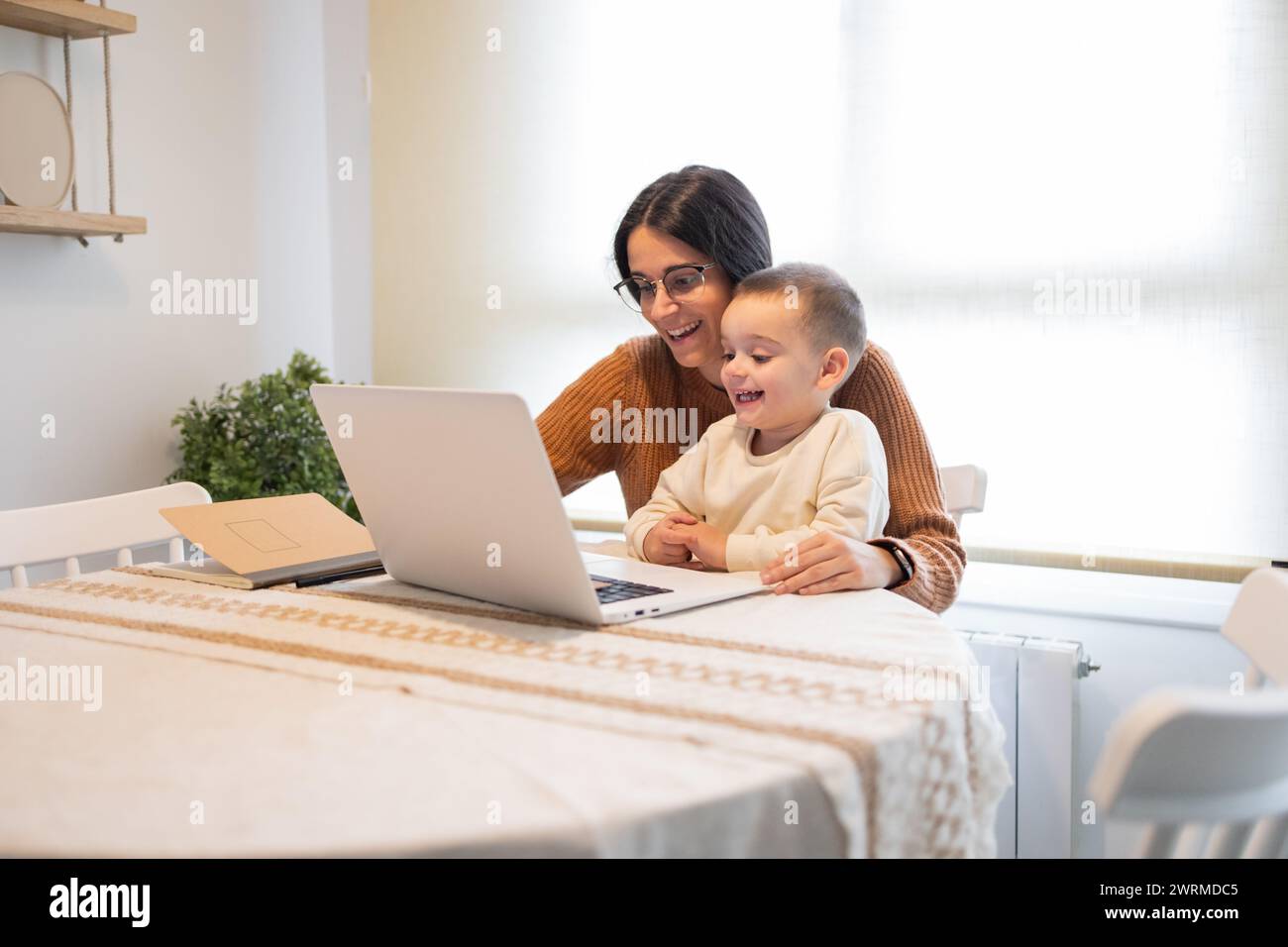 A heartwarming moment as a smiling mother embraces her son while they both engage with content on a laptop at home Stock Photo
