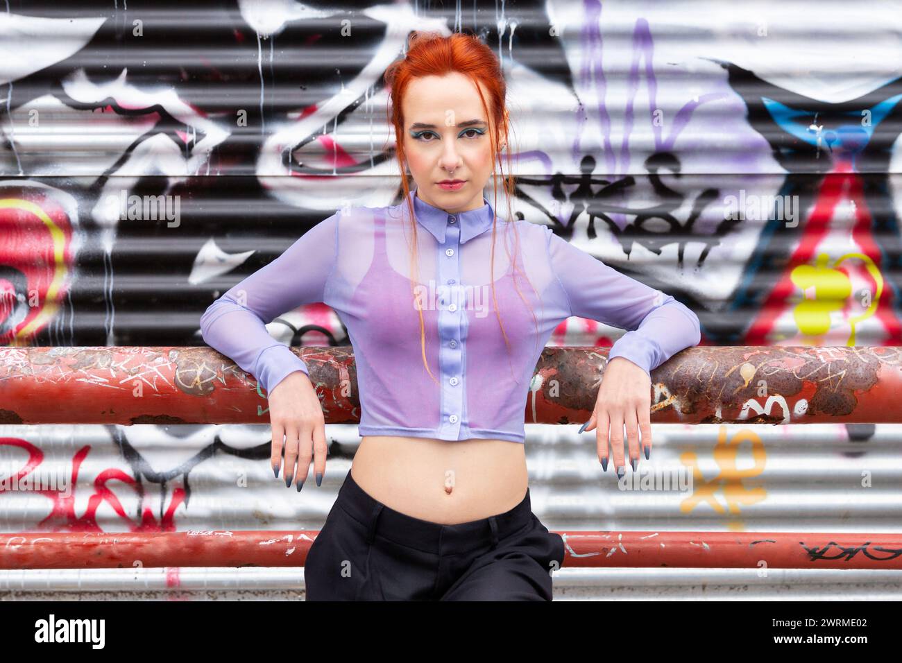 A Caucasian female poses against an edgy backdrop of colorful graffiti. Her striking red hair contrasts with her sheer lilac blouse and black slacks l Stock Photo