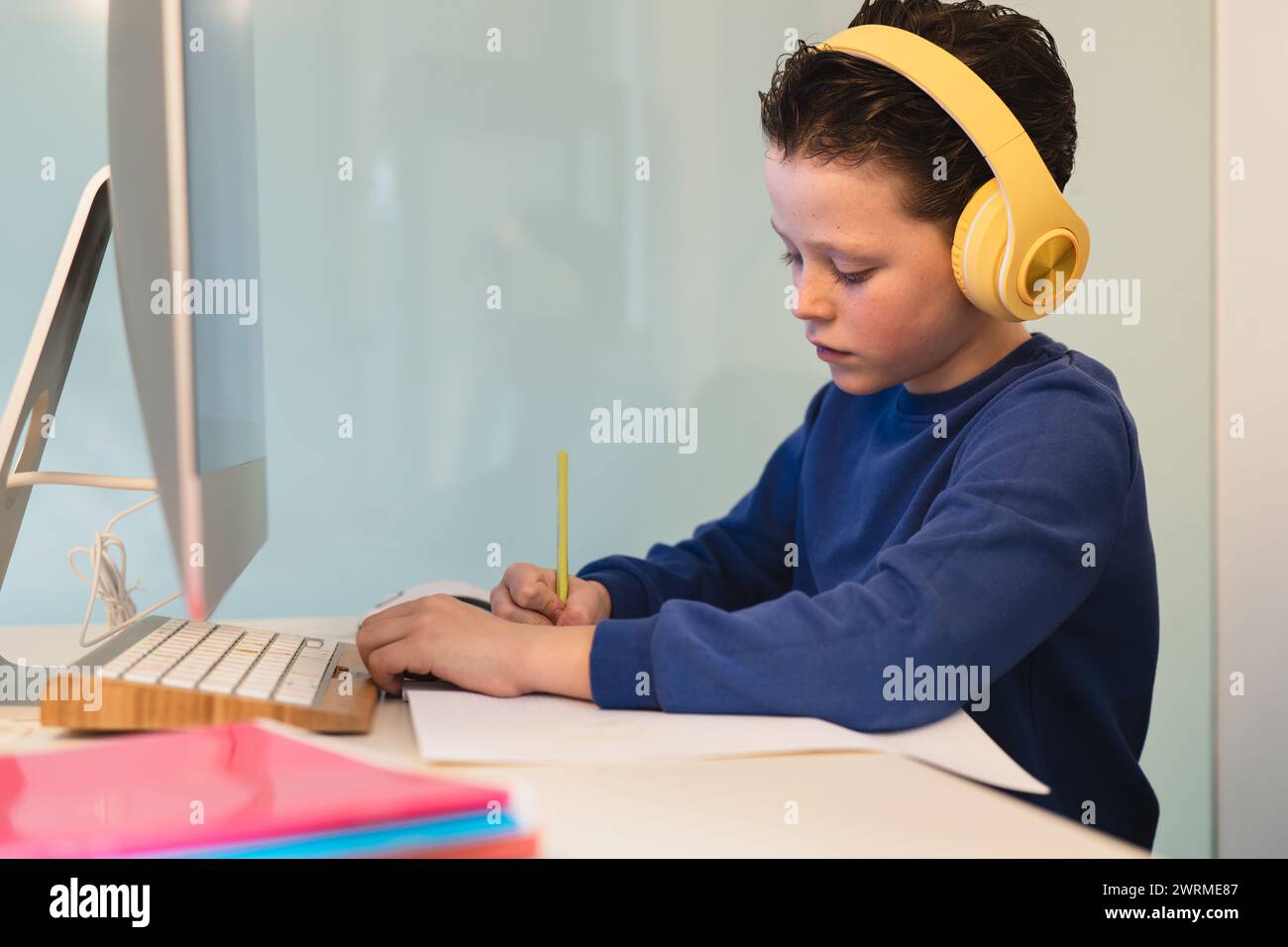 A dedicated child wearing headphones while writing notes at a desk with a computer, illustrating a modern multi-tasking study environment Stock Photo