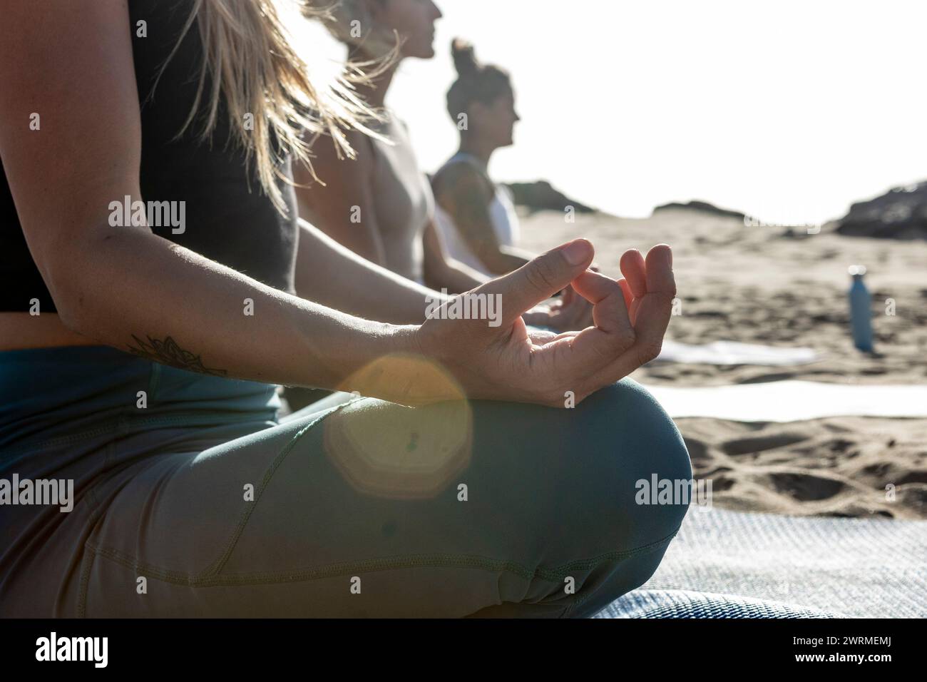 women participate in a tranquil yoga class on the beach, performing meditation with the sun setting in the background. Stock Photo