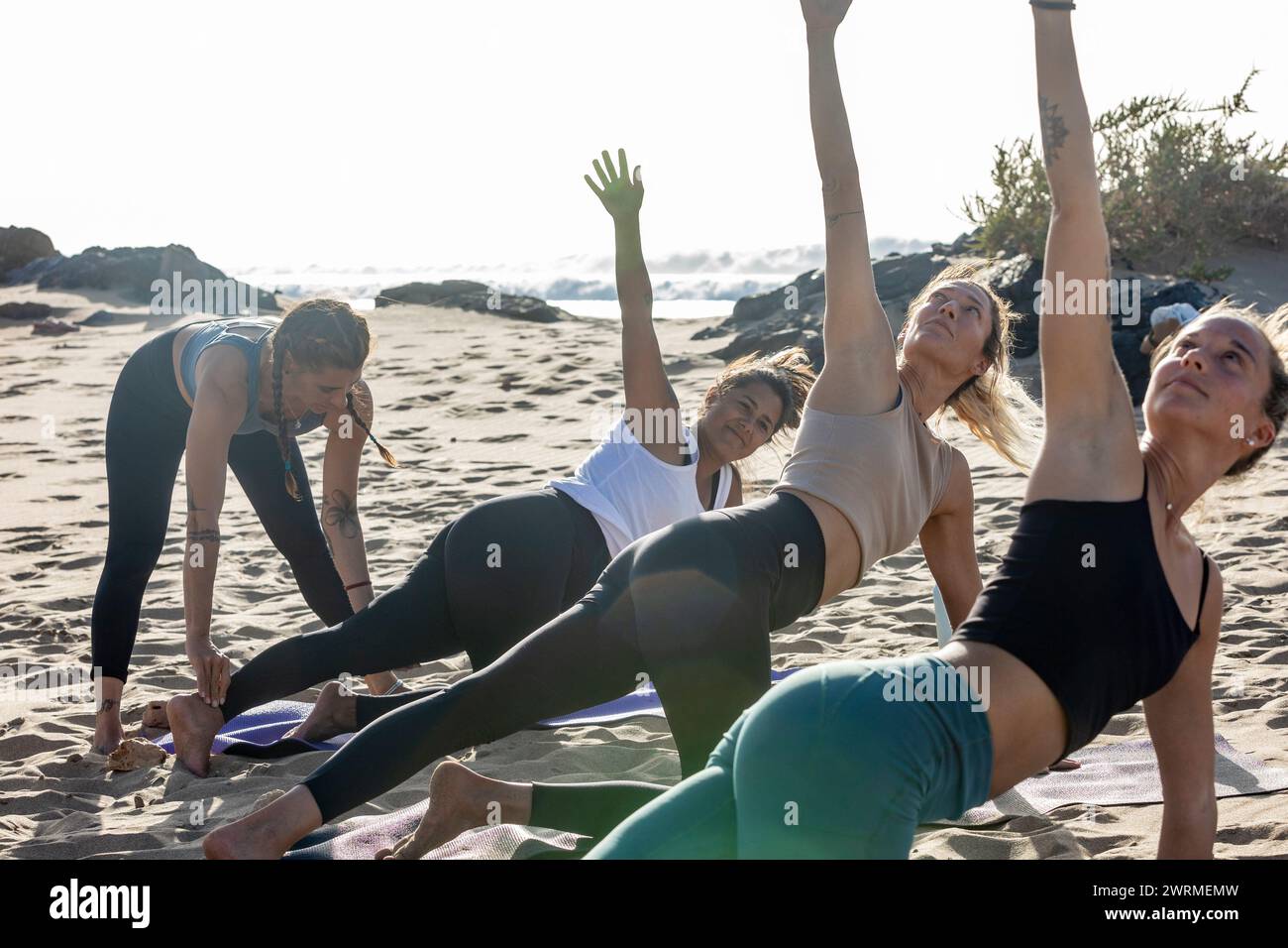 A group of women participate in a yoga class on the beach during sunset, with one person performing a Trikonasana pose. Stock Photo