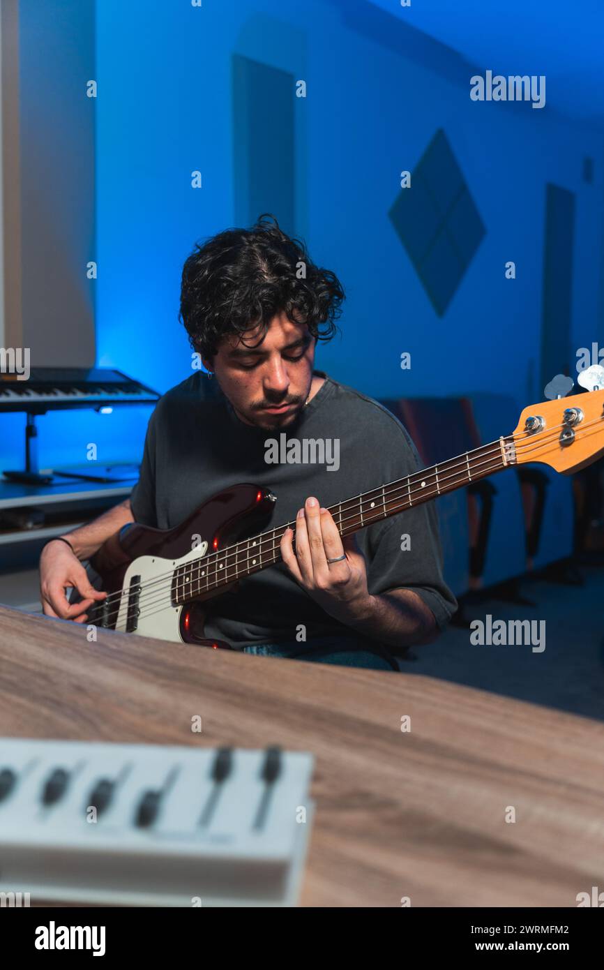 A focused male musician expertly plays a bass guitar during a sound studio recording session, with music equipment in the background. Stock Photo