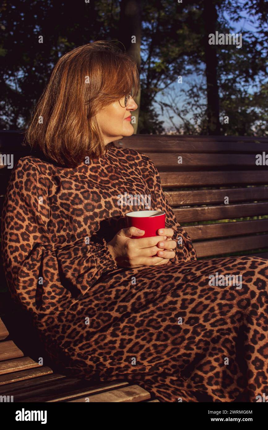 Woman in leopard dress with red cup. Woman in glasses smiling in park. Big city lifestyle. Morning coffee concept. Beautiful woman in sunlight. Stock Photo