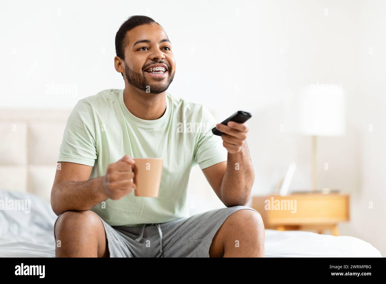Happy african american man sitting on bed, watching TV Stock Photo