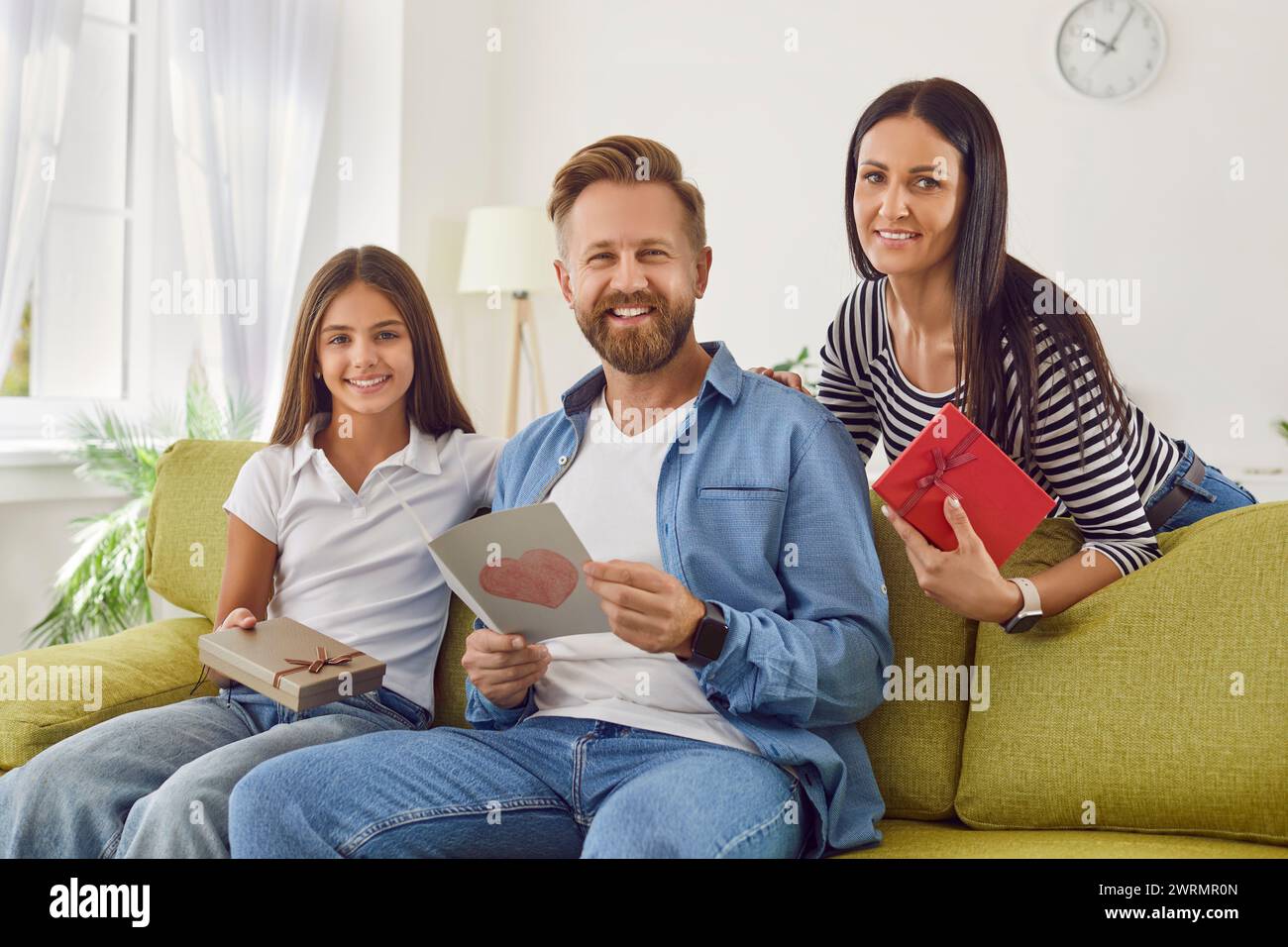 Father sitting on sofa at home with his girl child and wife and holding handmade greeting card. Stock Photo
