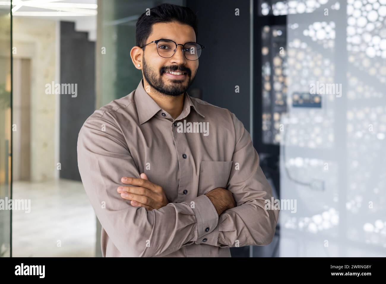 A professional young man with a beard and glasses, smiling confidently in a well-lit, contemporary office setting. Stock Photo